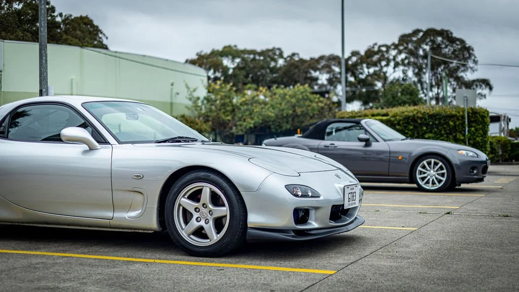 Two sports cars, a silver one in the foreground and a dark gray one in the background, parked in a parking lot with trees and a green building nearby.