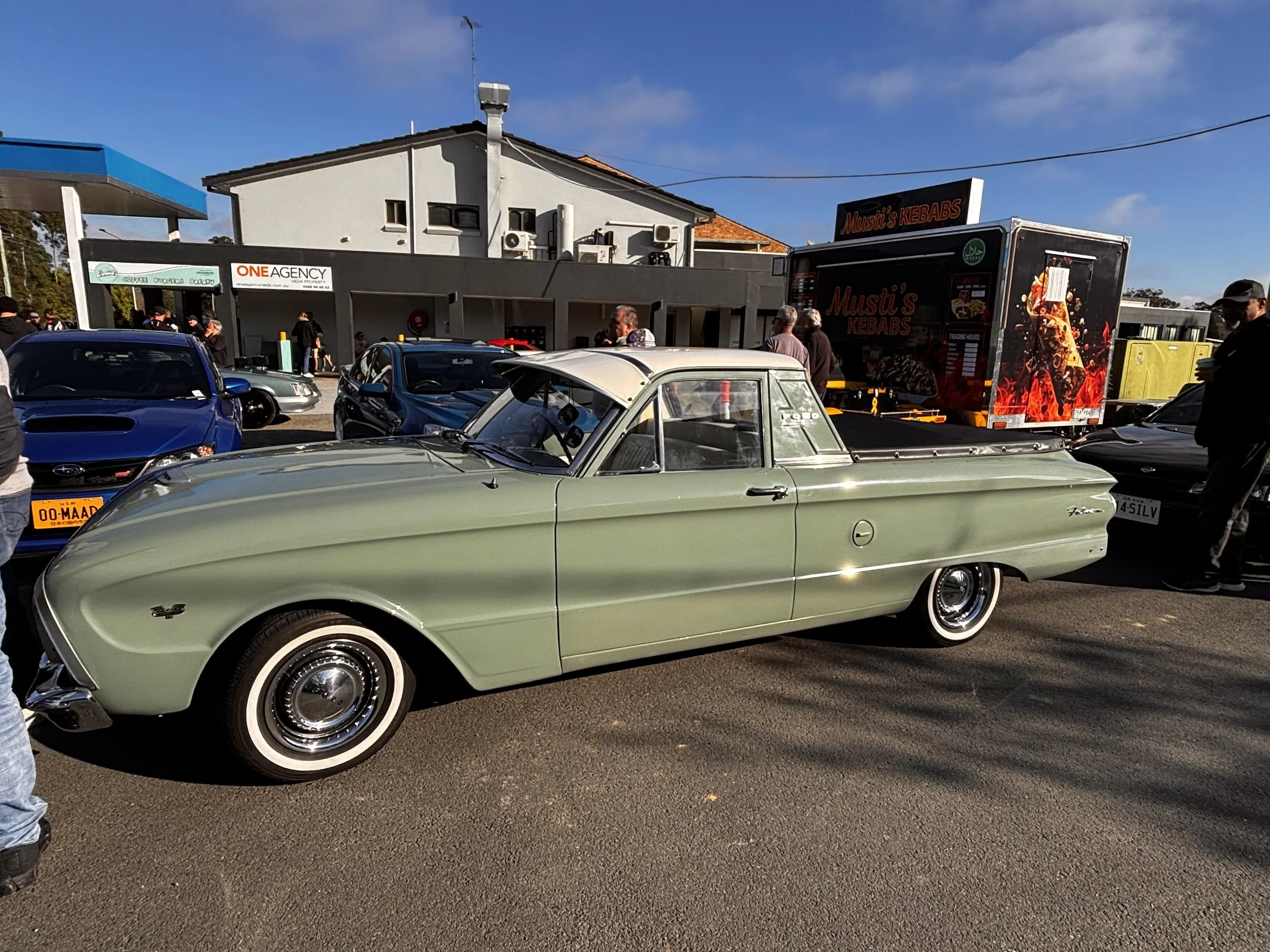A parking lot filled with vintage and modern cars, including a light green classic car in the foreground. In the background, there is a grey building with some air conditioning units and a sign for Mustang's Kebabs. The sky is partly cloudy with a br