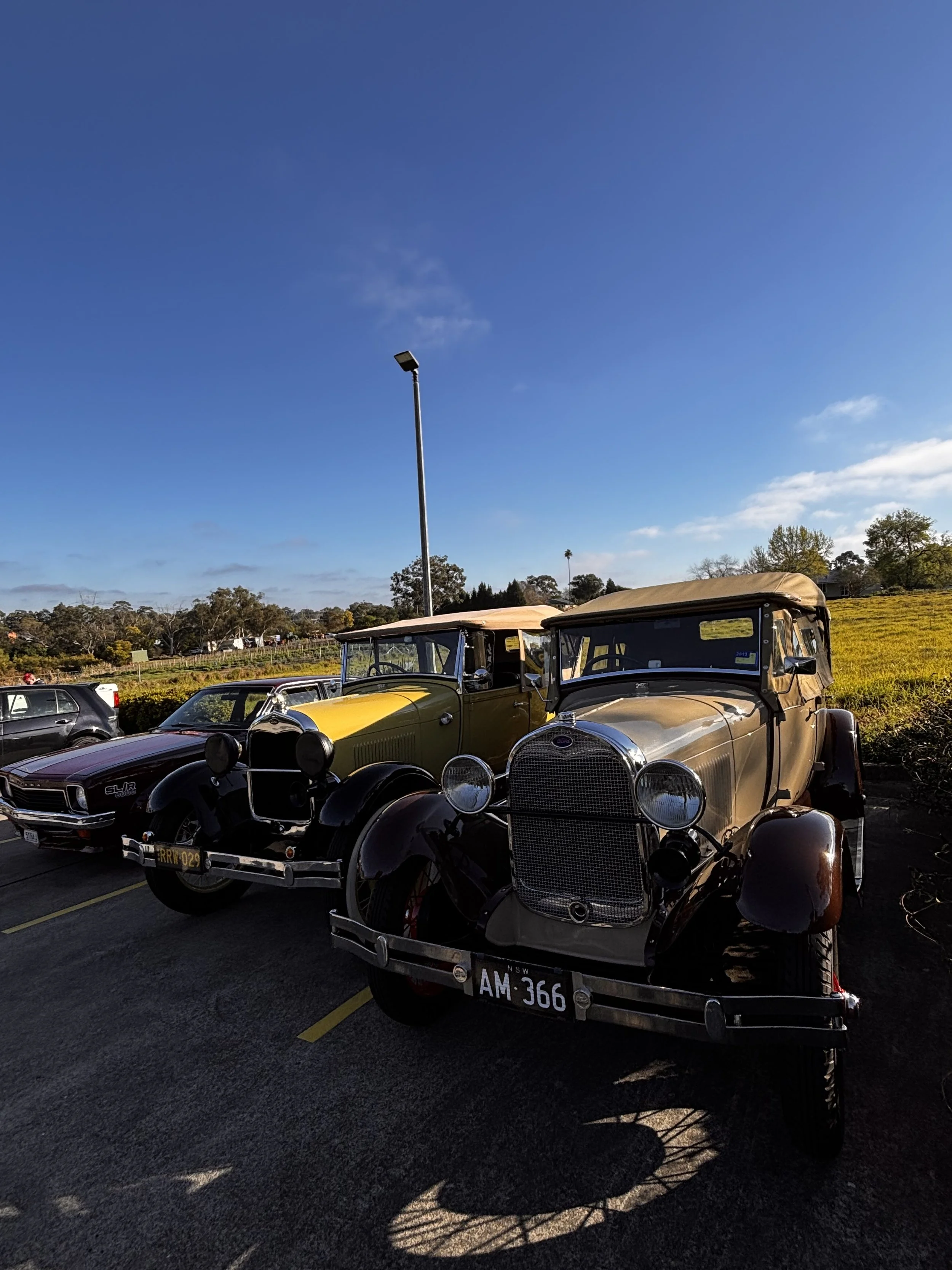 Several vintage cars parked in a lot under a blue sky with some clouds, including a black car and a tan-colored classic car in the foreground.