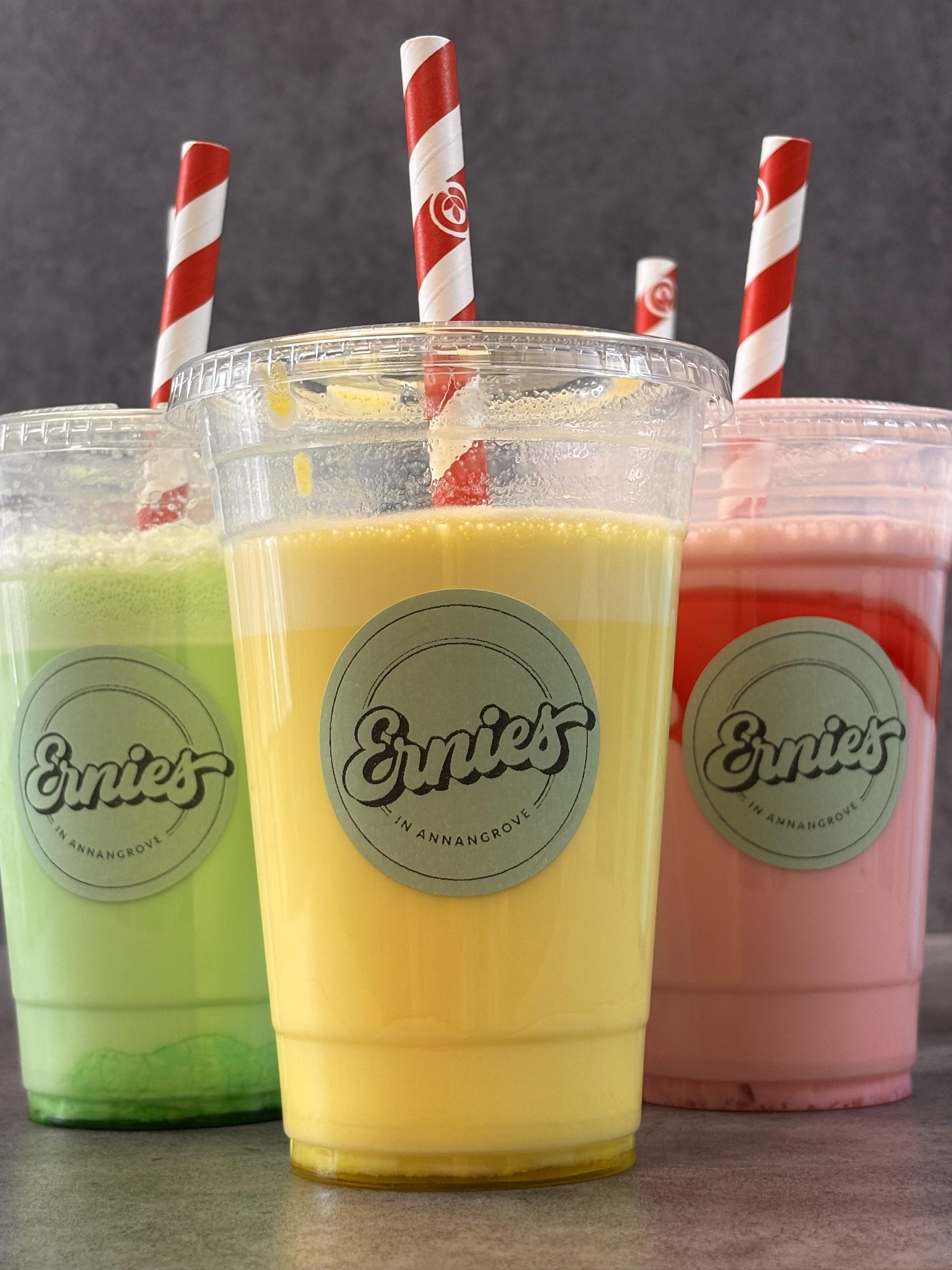 Three colorful smoothies in plastic cups with red and white striped straws, placed on a wooden surface, with a dark background. The cups have a label that says 'Emie's' and 'In Annangrove'.