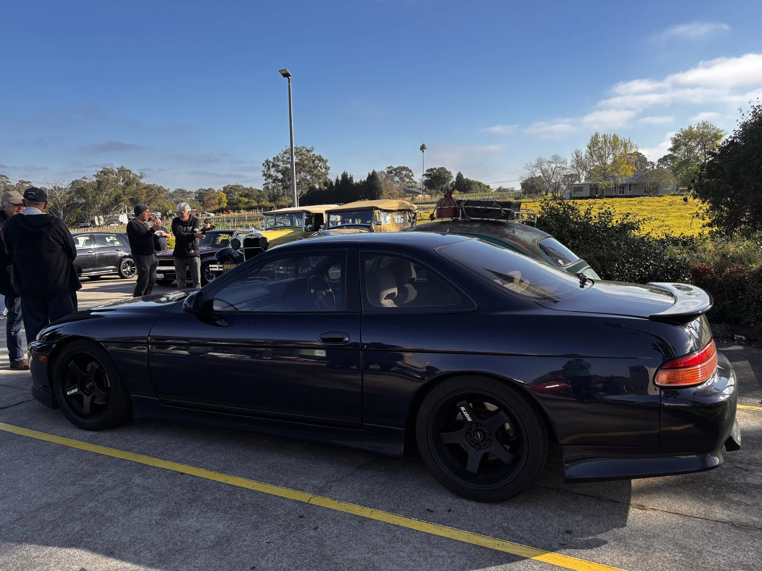 A parking lot filled with different cars and vintage vehicles, with a group of people standing and talking. In the background is a lush landscape with trees, a house, and a blue sky with some clouds.
