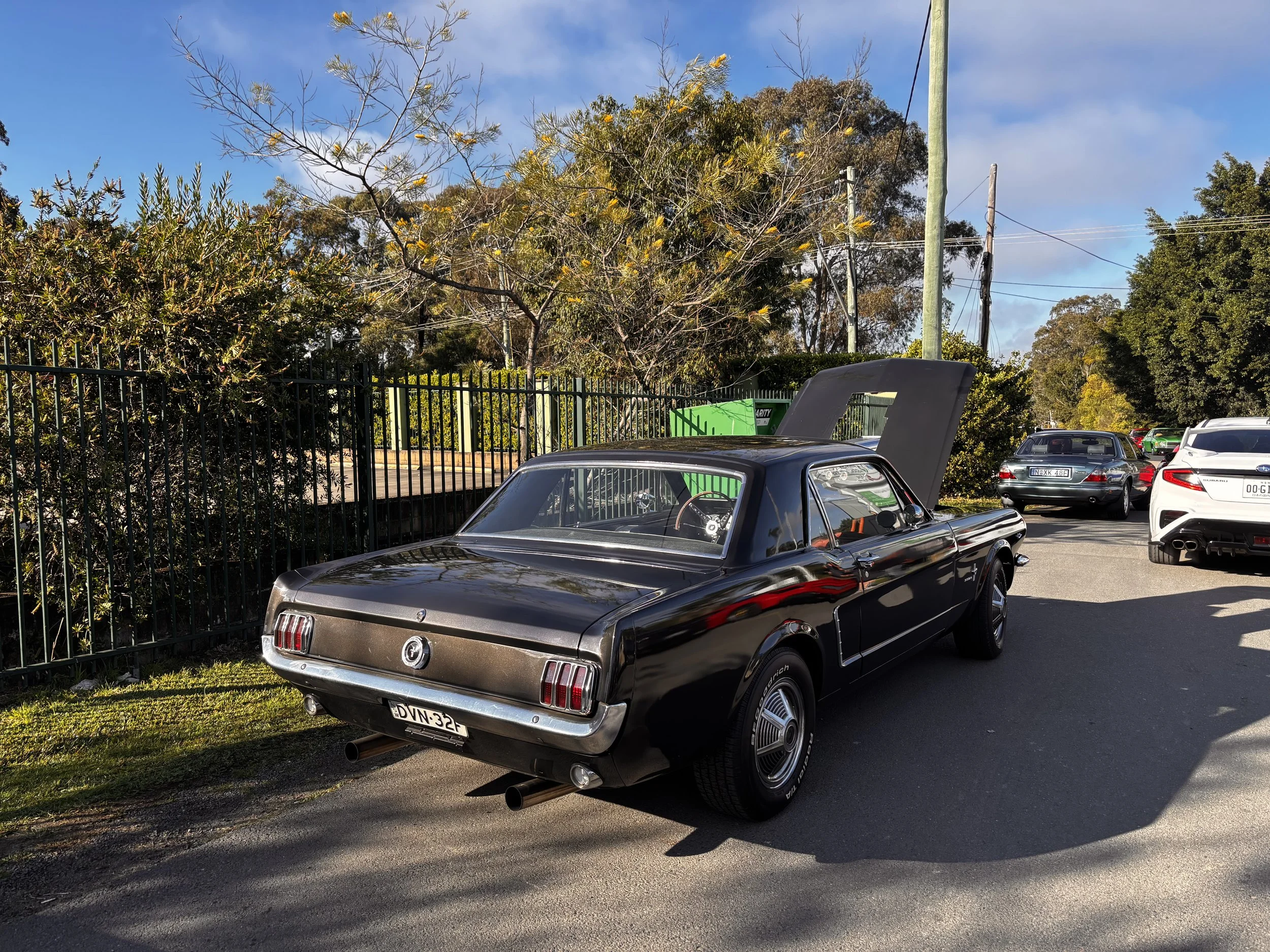 Black classic car parked in a parking lot with other vehicles, surrounded by trees and power lines, under a partly cloudy sky.