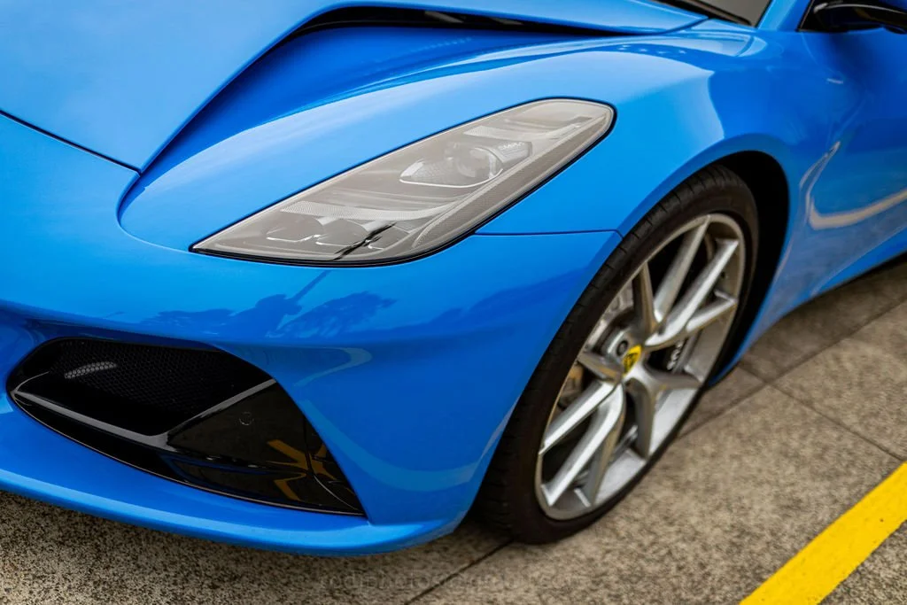 Close-up of a blue sports car, showing the front left side including the headlight, wheel, and part of the bumper, parked on a concrete surface.