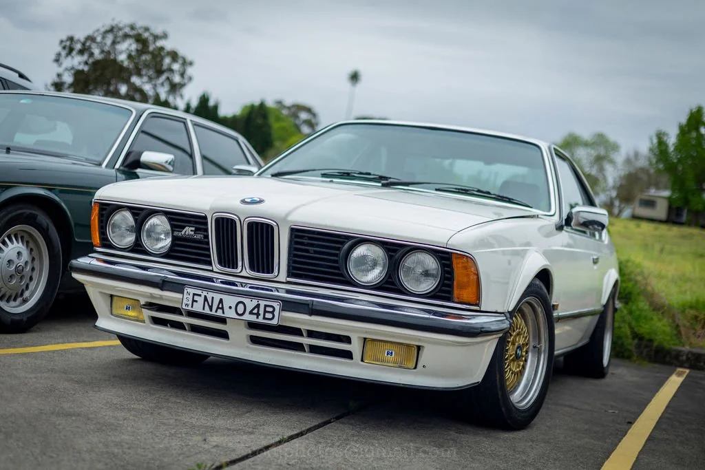 A white vintage BMW coupe with gold wheels parked in a parking lot, with another classic car beside it and greenery in the background.