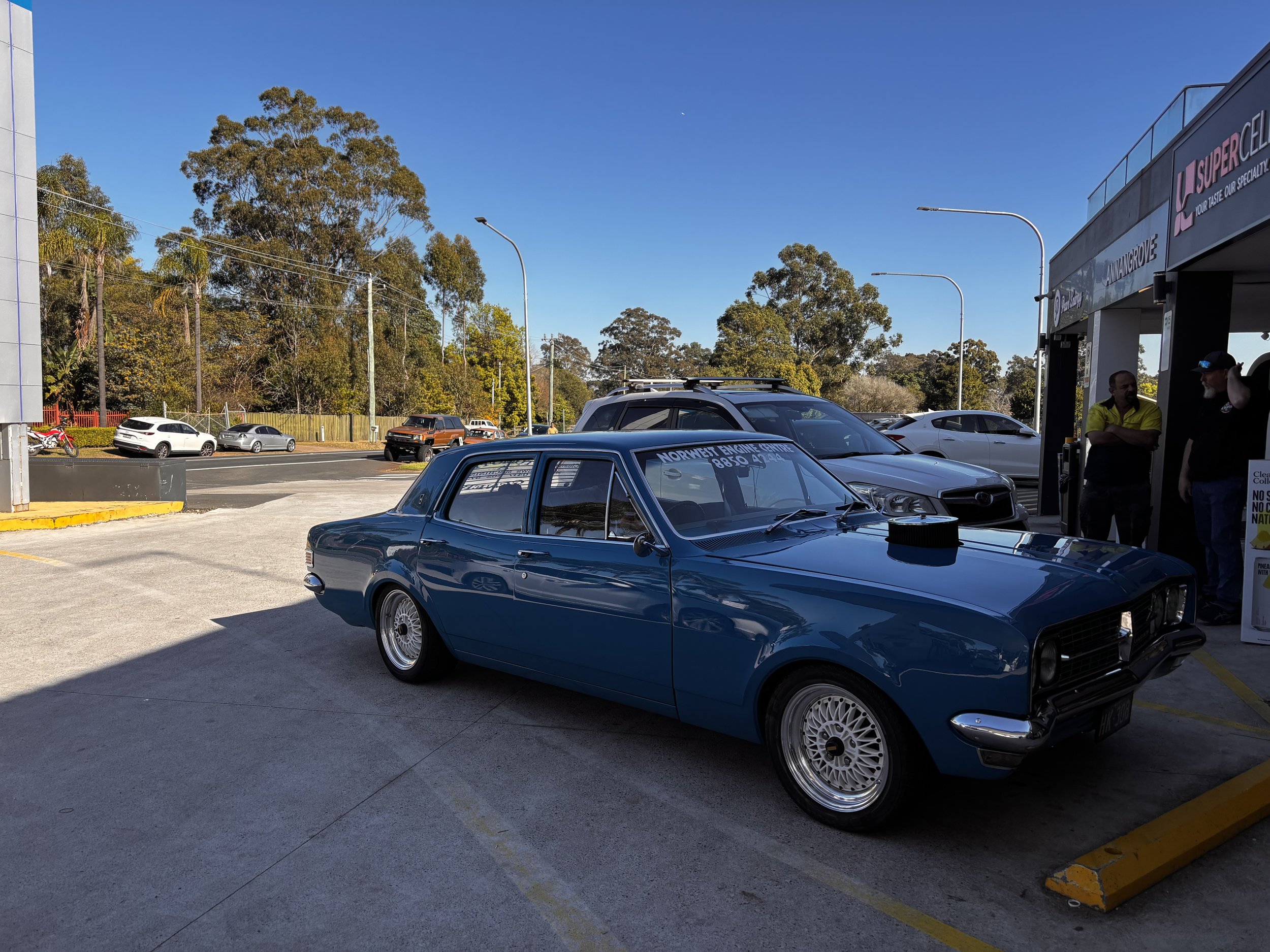 A parking lot outside a shopping center with several parked cars, some trees in the background, and a clear blue sky.