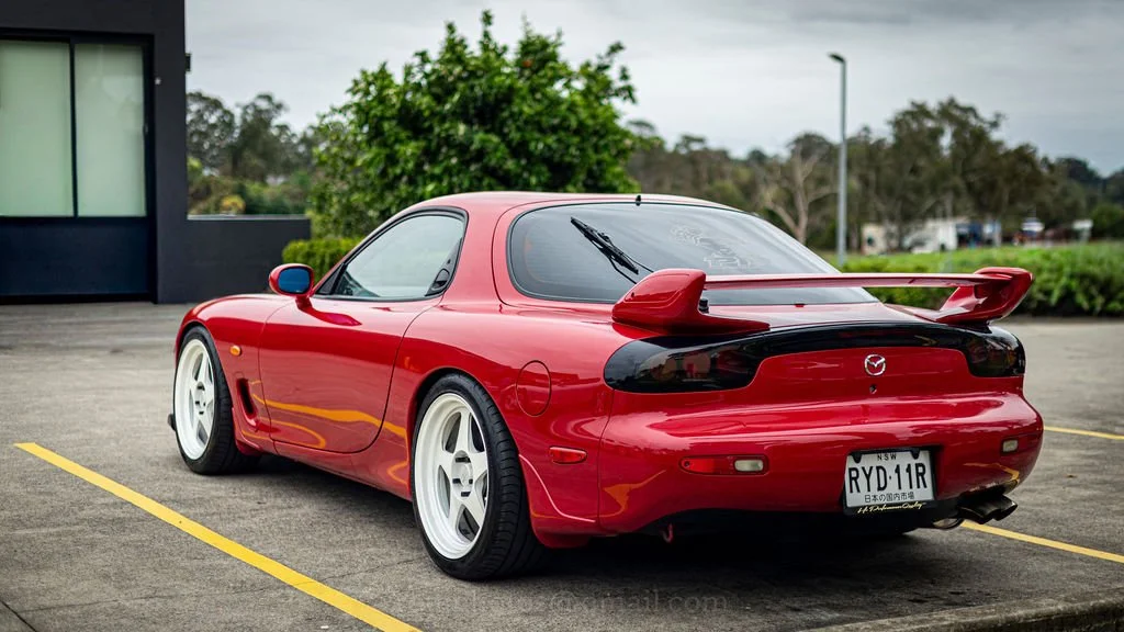 Red Mazda sports car with rear spoiler parked in an outdoor lot on an overcast day.