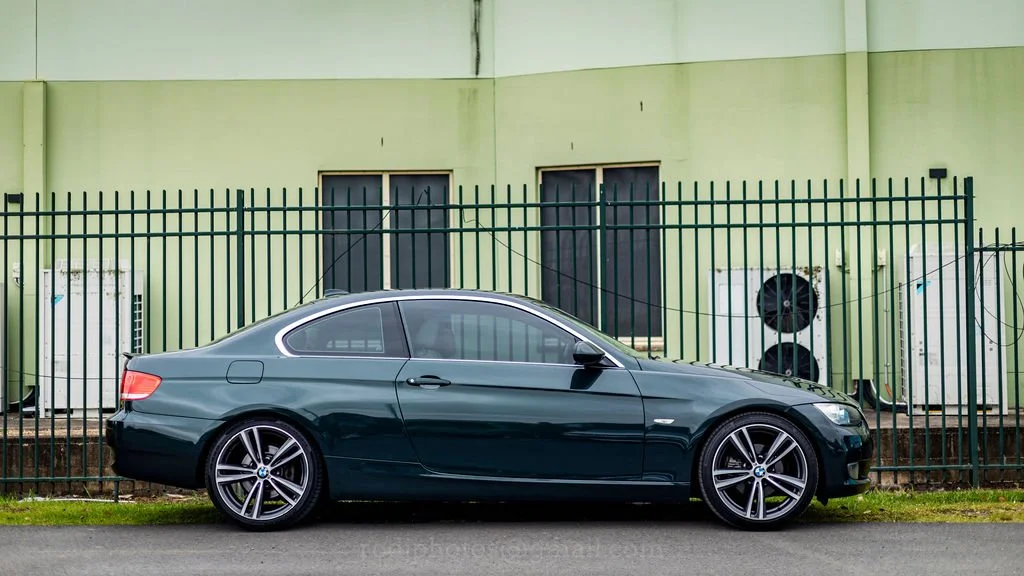 A dark green BMW parked beside a green building with a tall black fence in front.