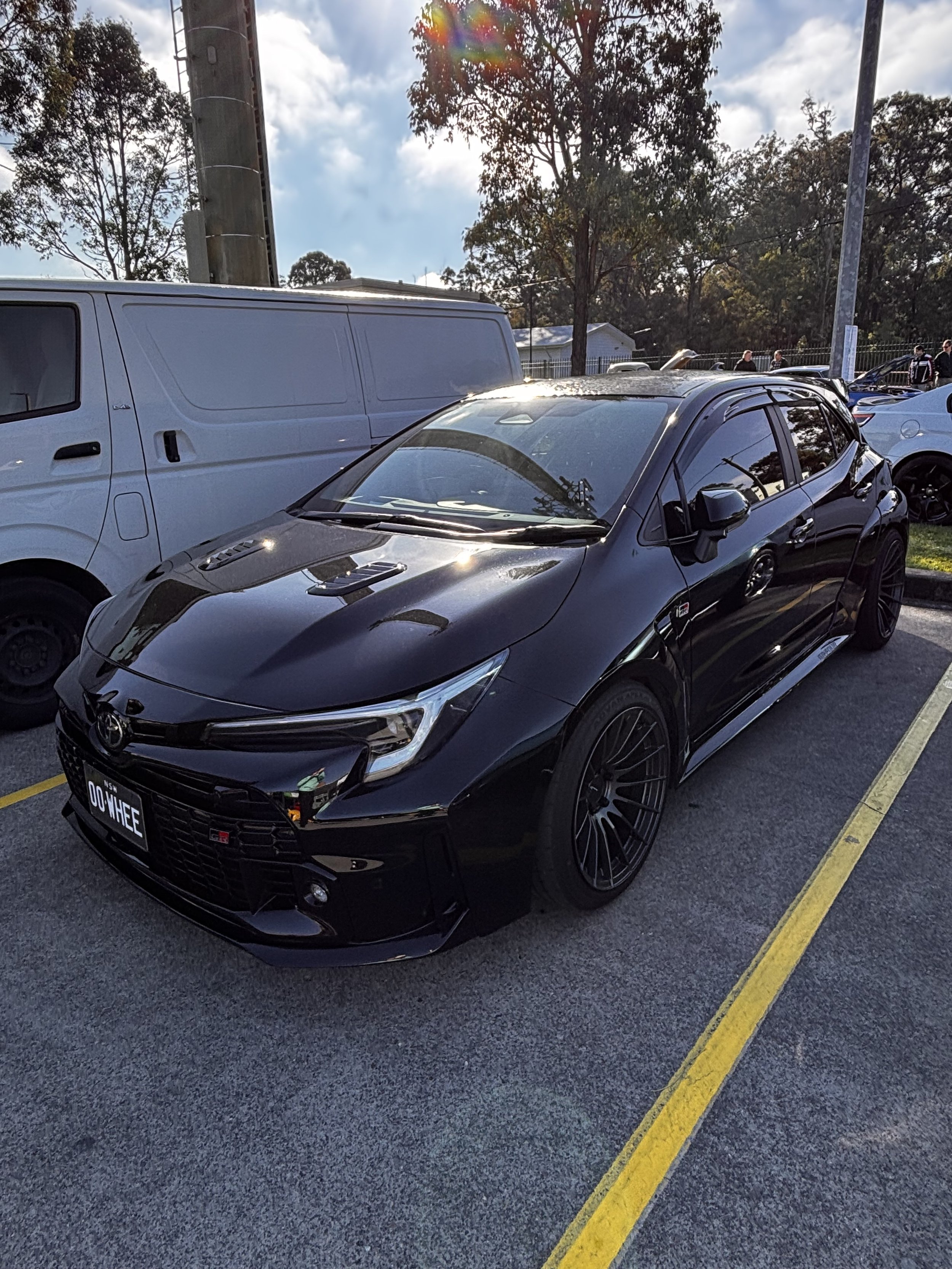 A black Toyota sports car parked in a parking lot with a white van behind it. The parking lot has a yellow line separating spaces, and there are trees, power poles, and a cloudy sky in the background.