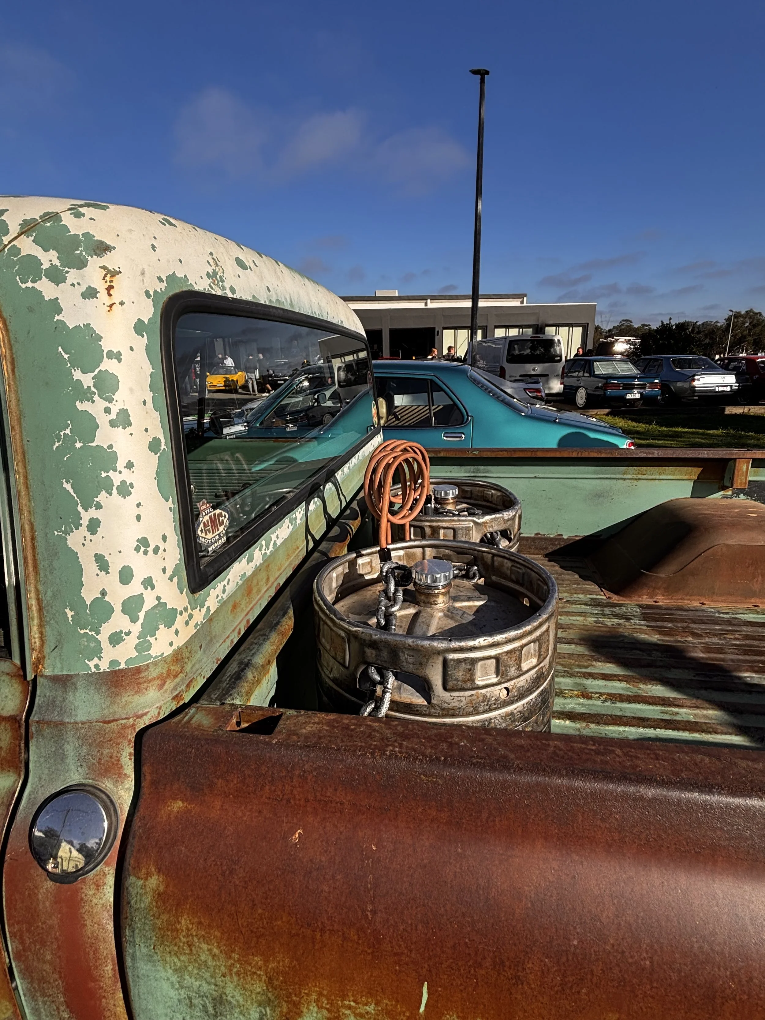 Close-up of an old, rusty pickup truck with a weathered, chipped green and rust-colored exterior. Inside, there are old barrels, one with orange hoses. Behind the truck, a parking lot with various cars and a modern building is visible under a bright 