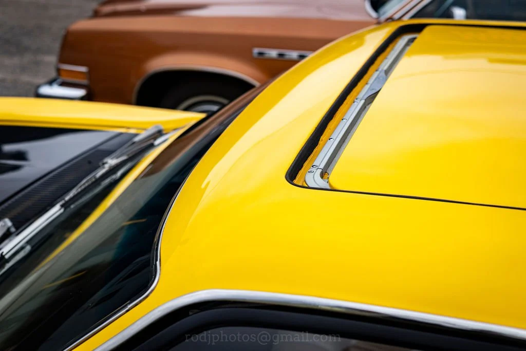 Close-up view of a yellow vintage car roof with a metal trim and black rubber weatherstripping, parked alongside a brown car in the background.