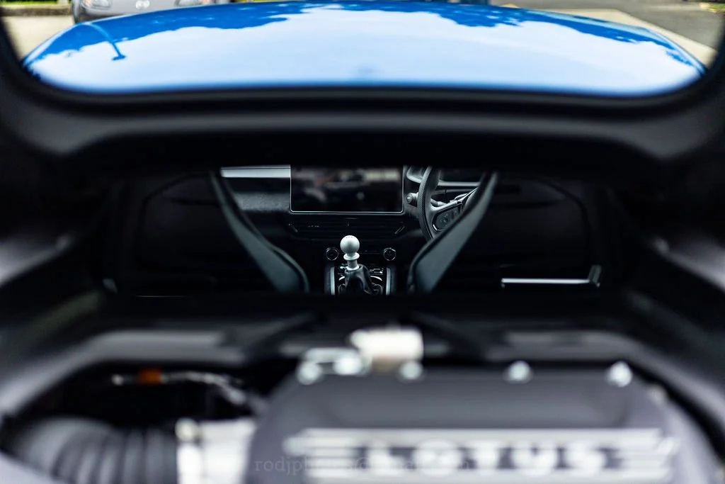 A view of a car's interior from the back seat looking forward, showing the dashboard, steering wheel, and a digital display screen, with part of the windshield and reflection of the sky.