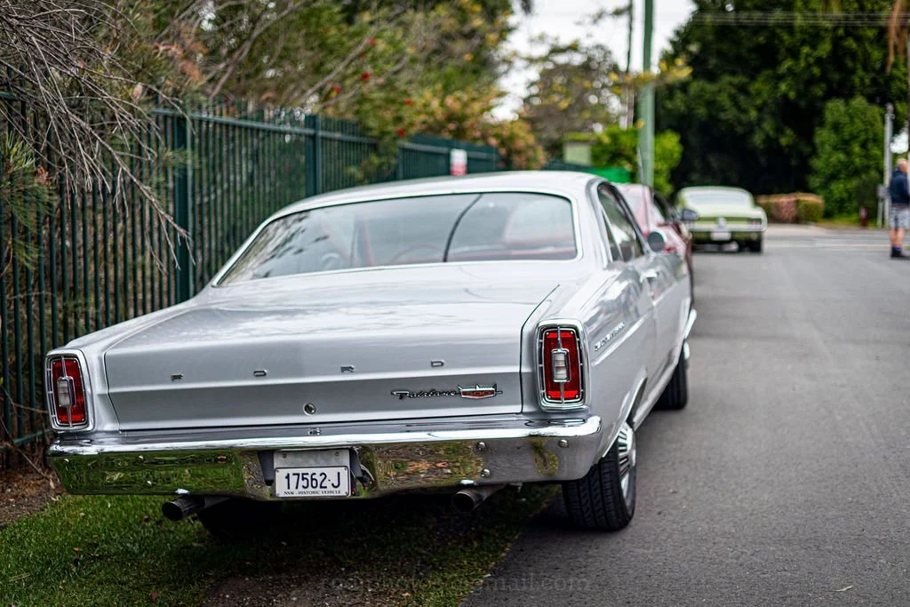 A vintage silver Ford Thunderbird parked on the side of a street with a green fence and trees in the background.