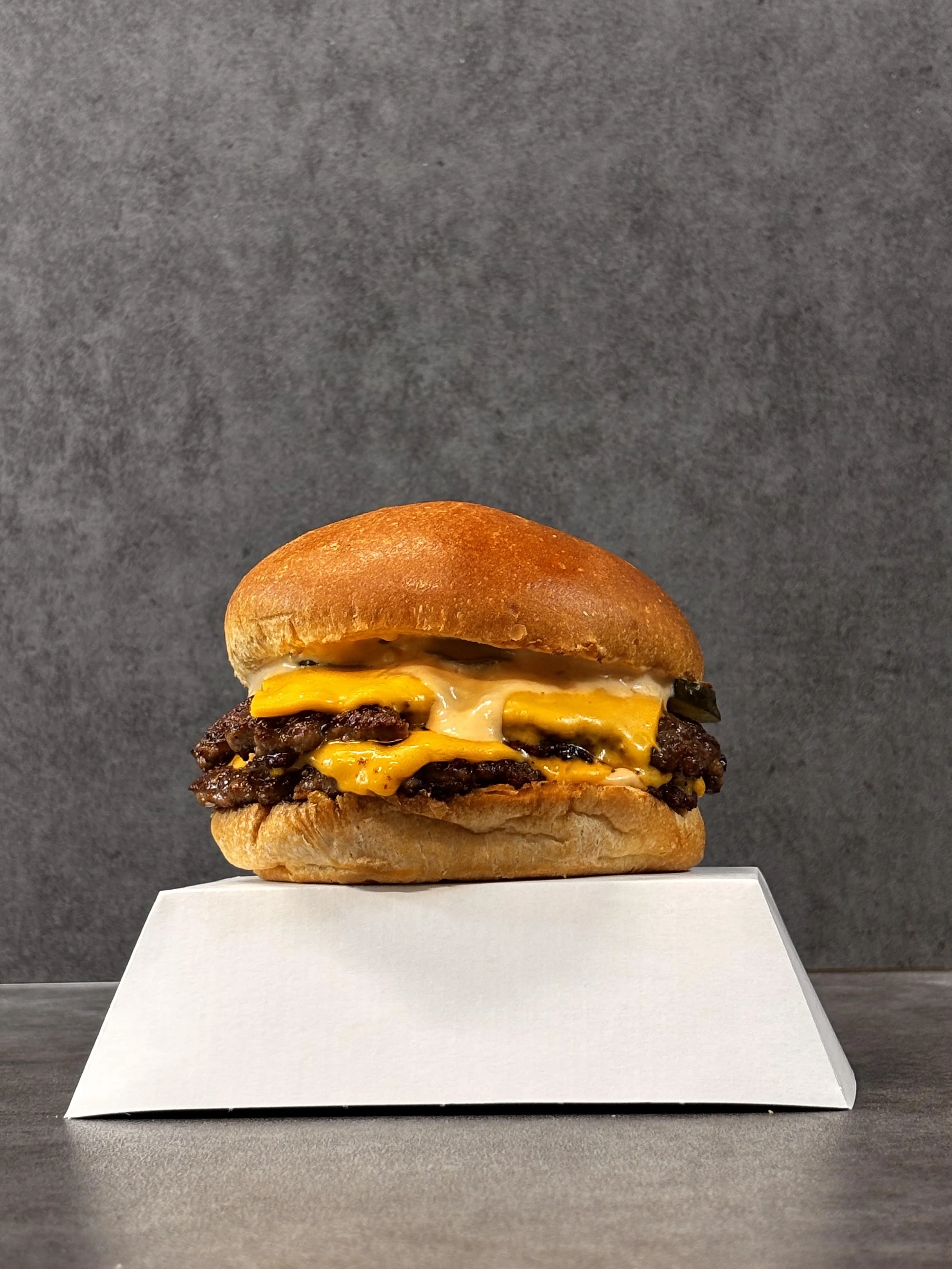 An assembled cheeseburger with cheese, beef patty, bun, and toppings on a white box against a dark gray background.
