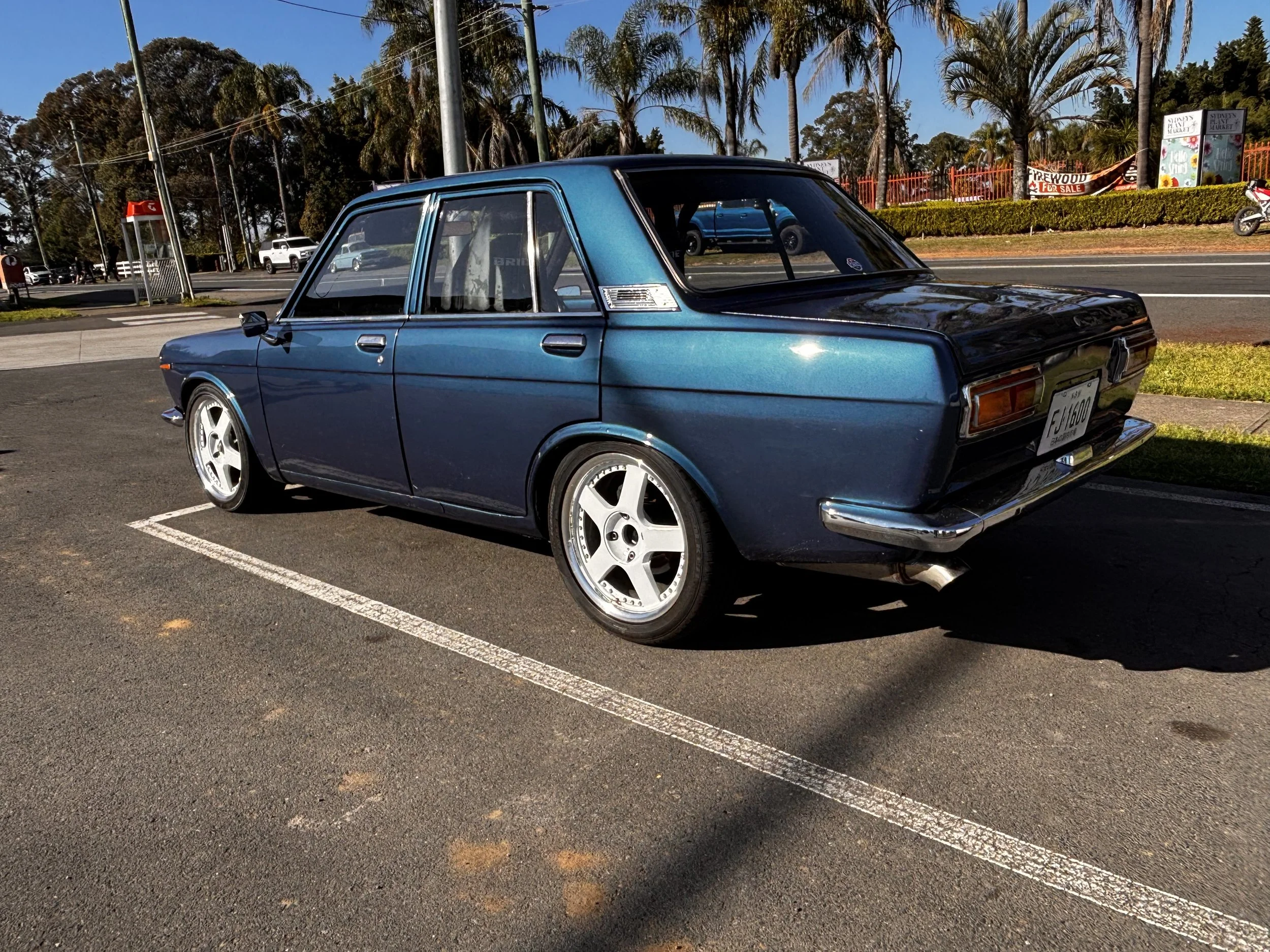 A vintage blue sedan car parked in a parking lot with a background of trees, palm trees, and a street. There are other vehicles and a motorcycle in the background along with a yard sale sign.