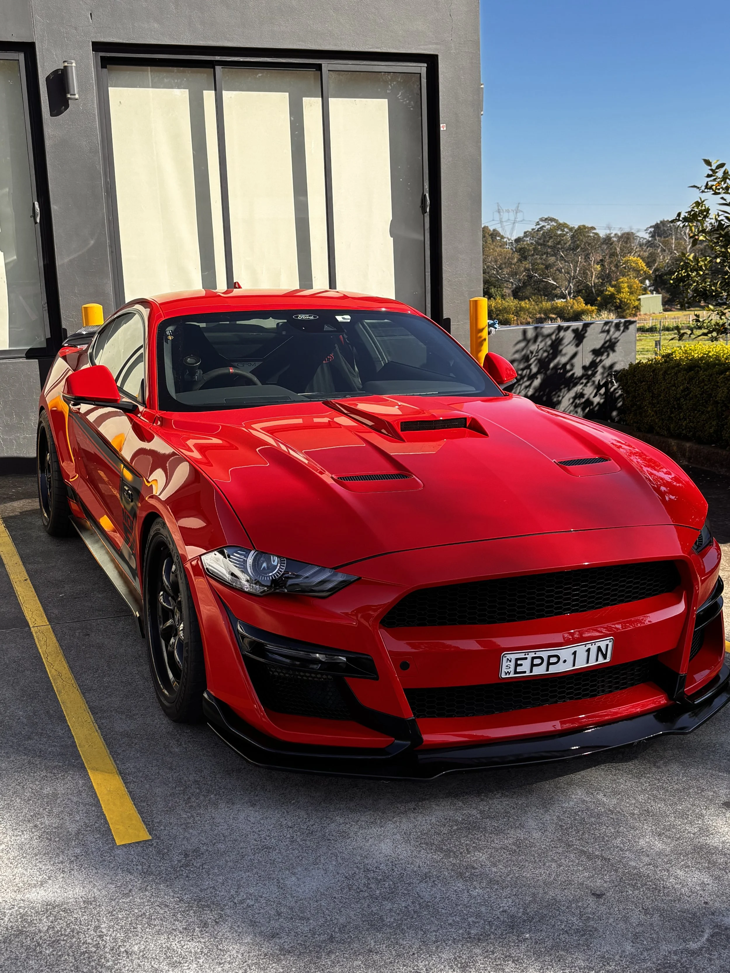 A red sports car parked in a parking lot next to a building with large windows. The car has a license plate from New South Wales, Australia, and features a sleek, aerodynamic design with a scoop on the hood.