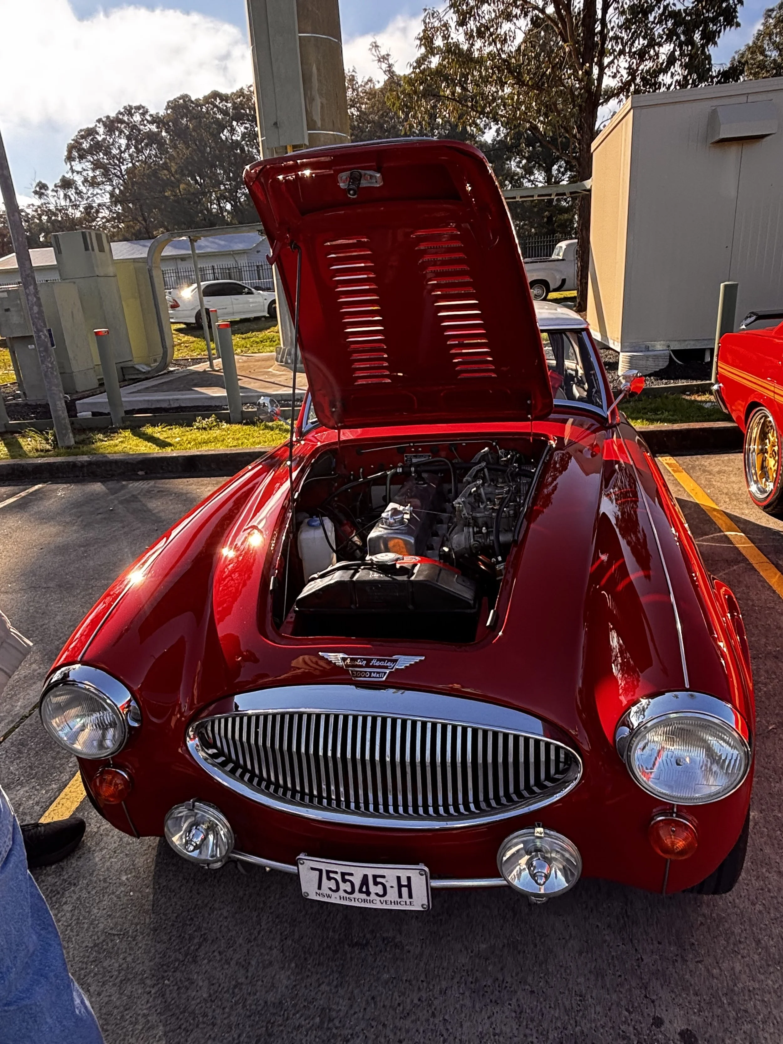 A red vintage racing car with its hood open, showing the engine inside, parked in a lot with other vehicles in the background.