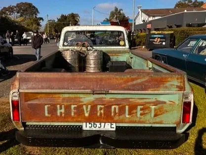 Rear view of a vintage Chevrolet pickup truck with a rusted tailgate parked on a street, with other cars and people in the background.