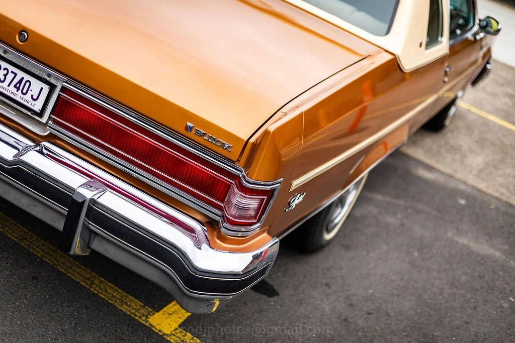 Rear side of a vintage orange Buick car with chrome bumper and taillight, parked in a parking lot.