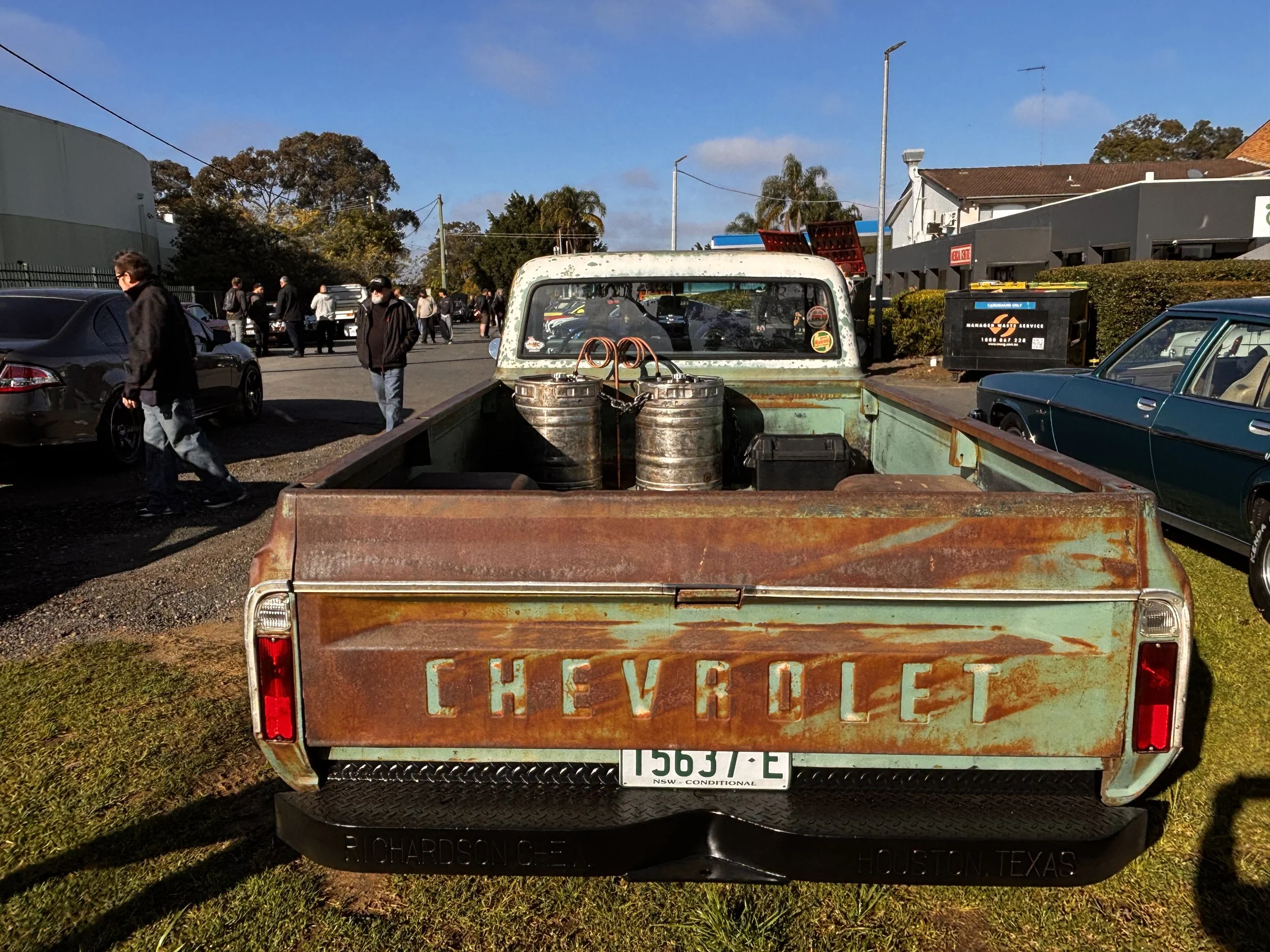 View of a parking lot with several cars and people walking, with a rusted Chevrolet truck in the foreground and a building with a sign in the background.