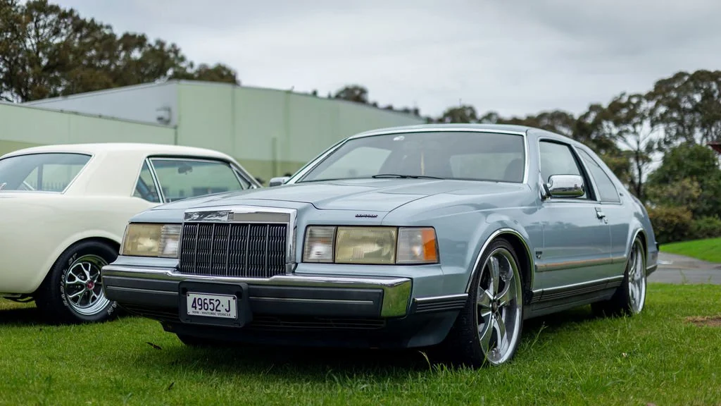 A vintage light blue two-door car with chrome accents, large custom wheels, and a black front grille, parked on a grassy area next to another classic white car.
