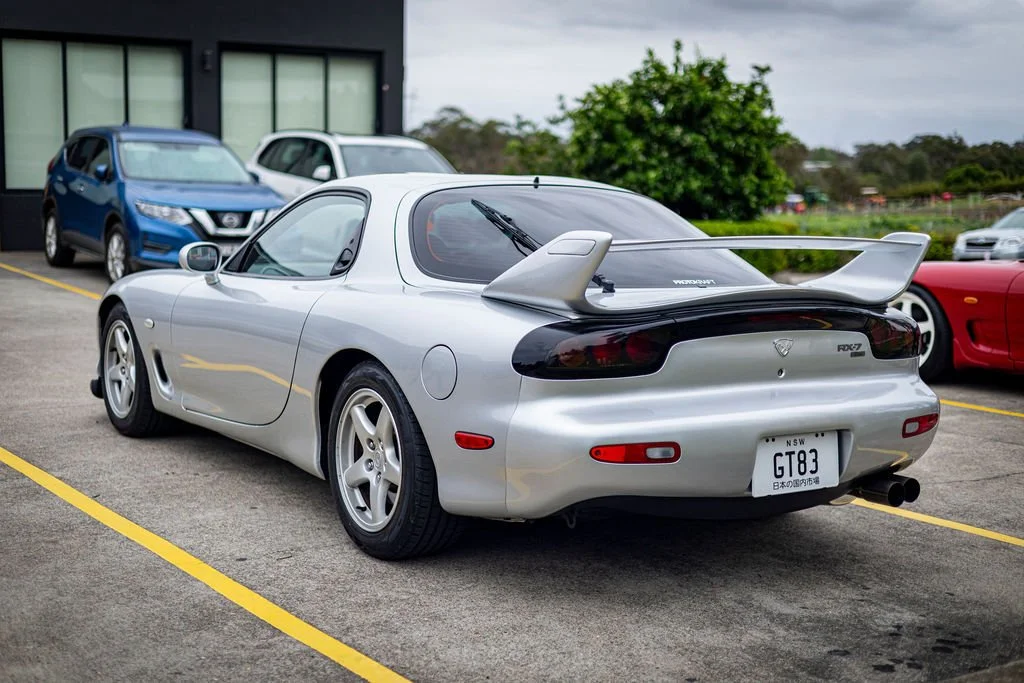 Silver Toyota Supra with a large rear wing, parked in a lot with other cars and trees in the background.
