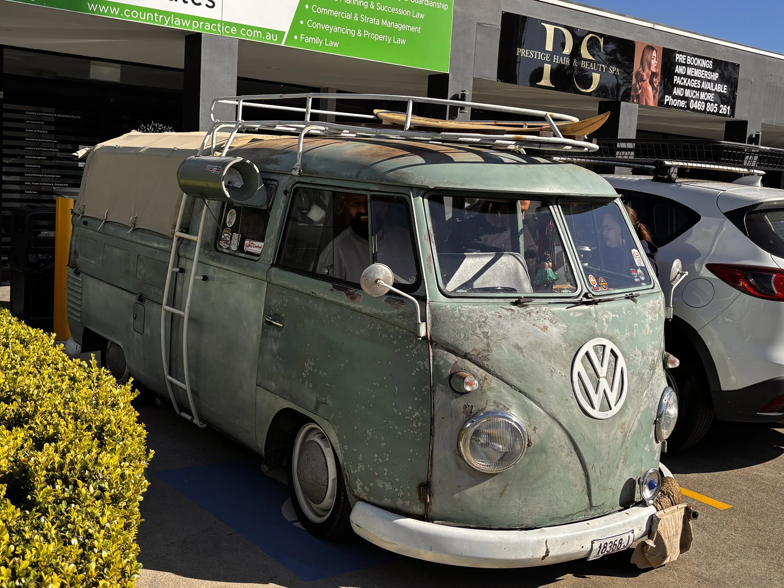 An old, rusty Volkswagen Van with surfboards on the roof rack parked in a parking lot near a building. The van's exterior is weathered, and the front bumper appears damaged.