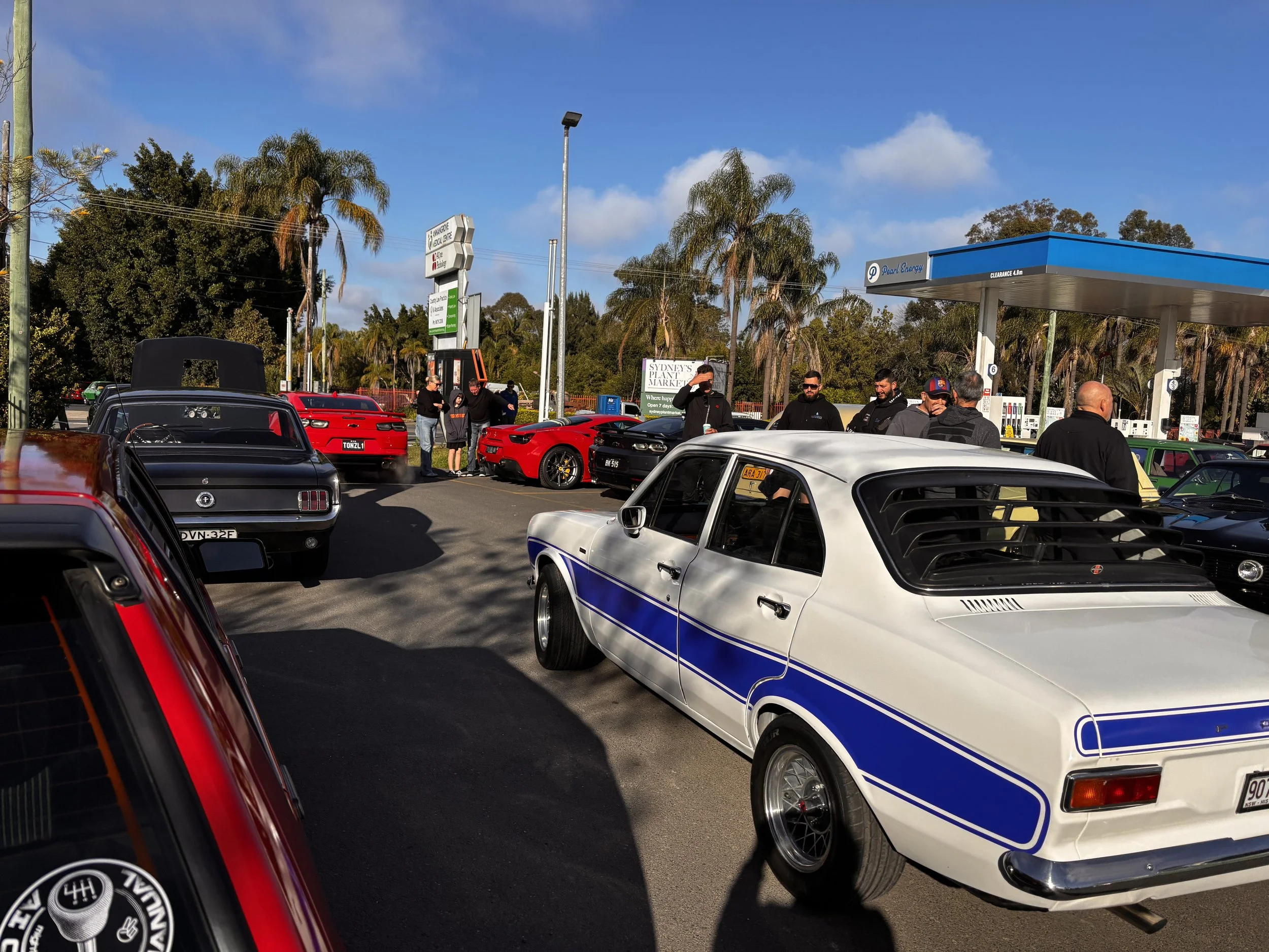 A parking lot with several classic and modern cars, people chatting, and a fuel station named Pearl Energy under a partly cloudy sky.
