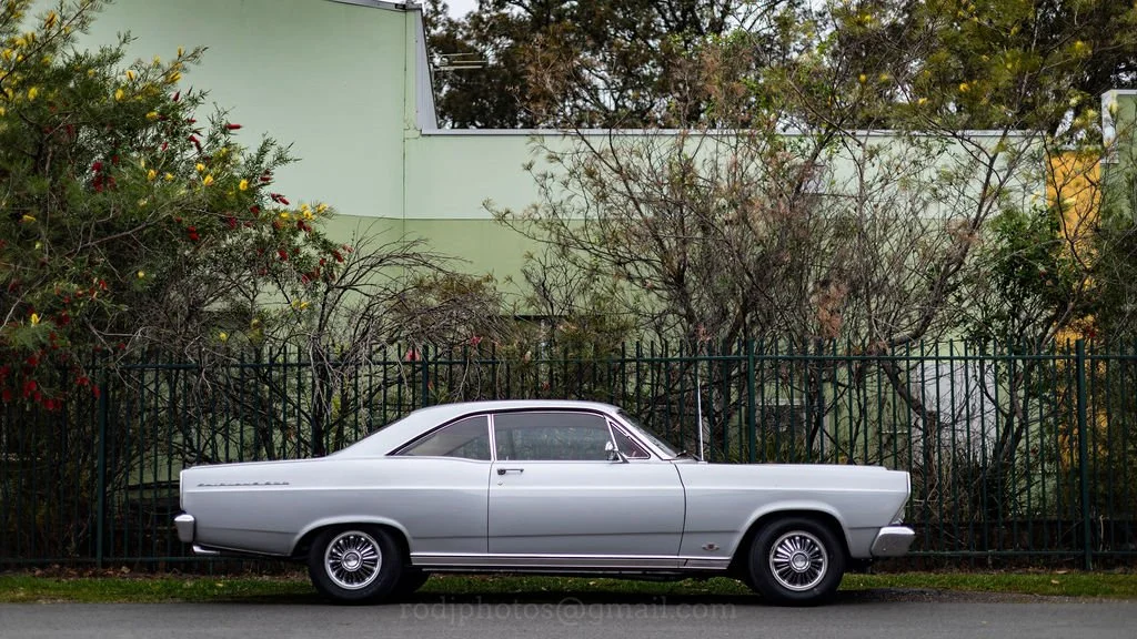 A silver vintage car parked on the street in front of a green building and a fence, with trees and bushes in the background.