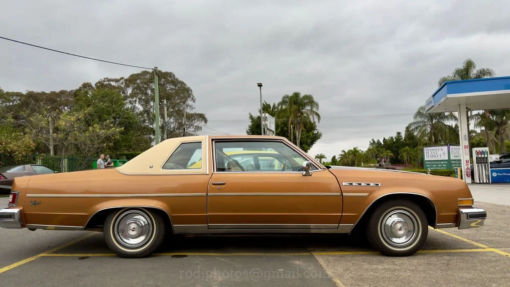 A vintage brown Oldsmobile Cutlass parked at a gas station with a blue roof, overcast sky, and palm trees in the background.