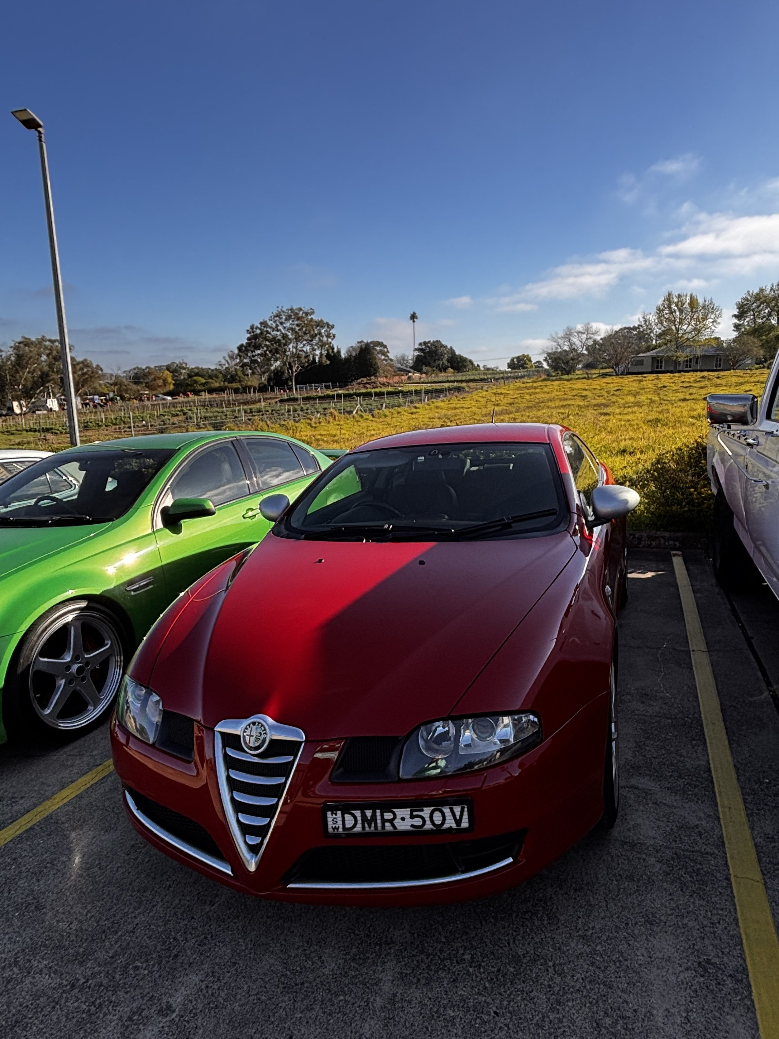 Red Alfa Romeo car parked in a parking lot next to a green car, with a field and trees in the background under a blue sky.
