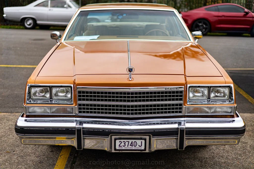 Front view of a vintage gold-colored Buick car parked in a parking lot with other vehicles in the background.