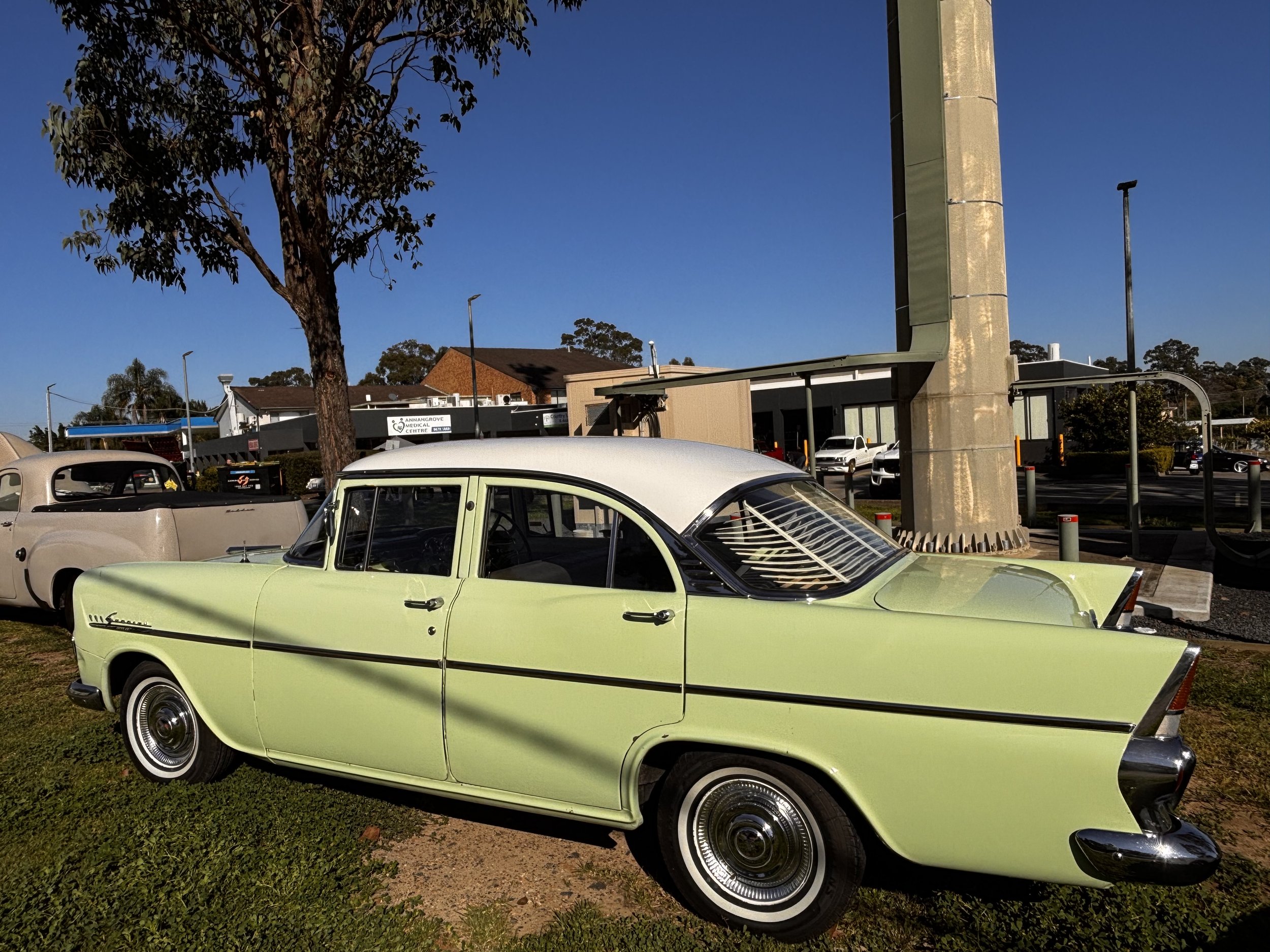 A light green vintage car parked on grass next to a concrete pillar, with other cars and buildings in the background and a tree on the left side of the image.