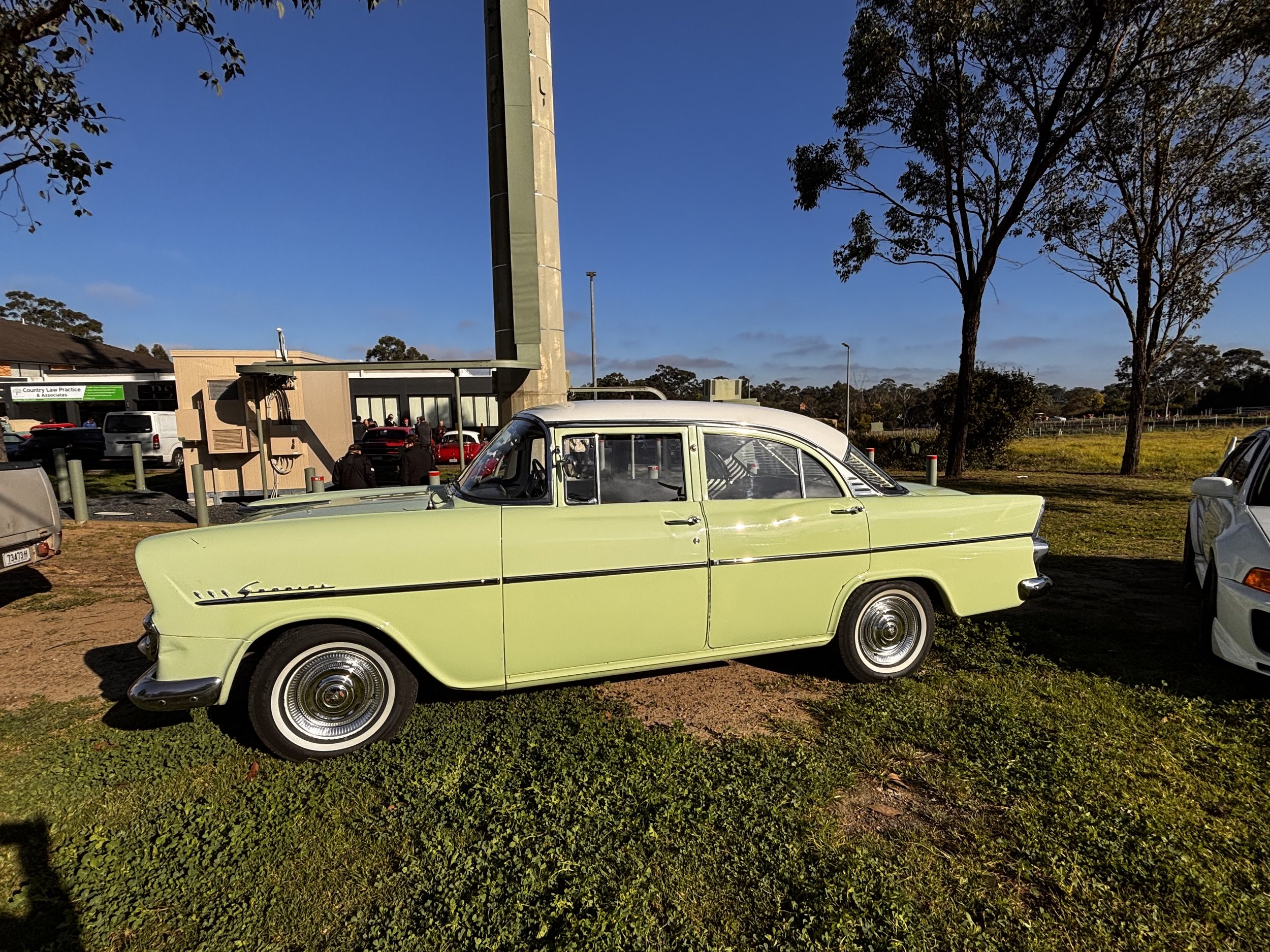 A parking lot with several parked cars, including a vintage light green car in the foreground. Behind the cars, there is a small building, some trees, and a clear blue sky.