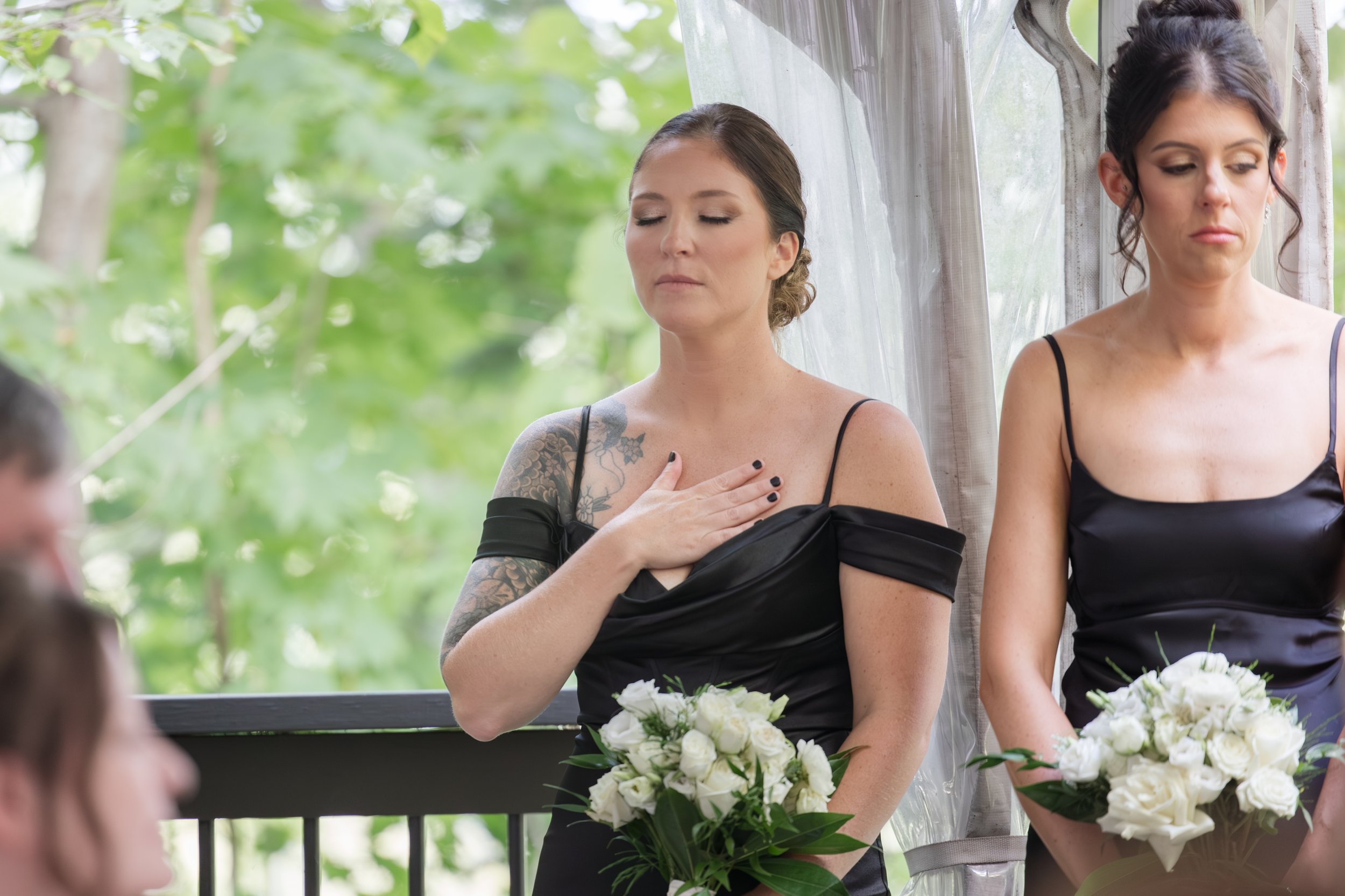 Two women in black dresses holding white flower bouquets at a wedding ceremony, with one woman placing her hand on her chest during a moment of reflection or prayer.