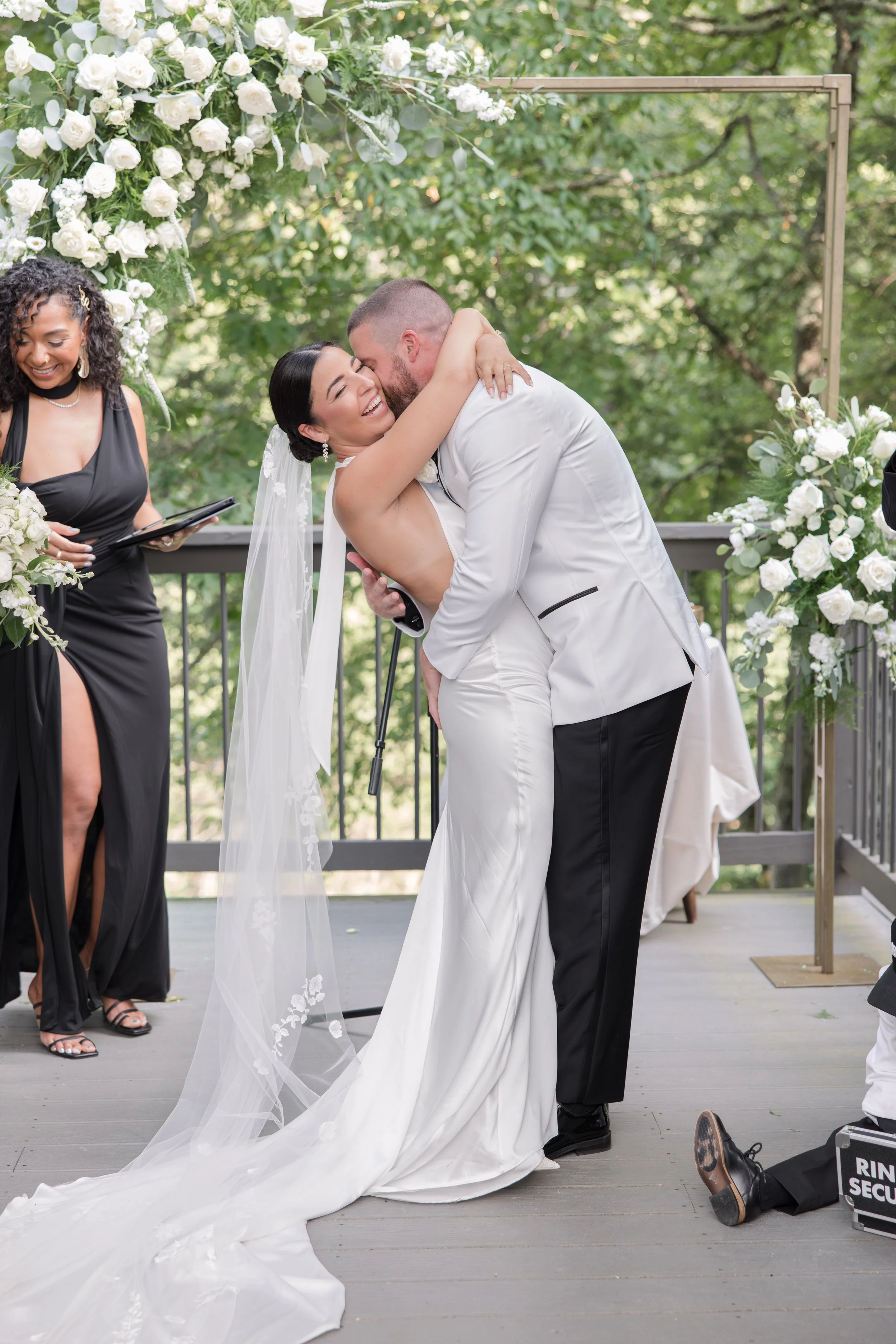A bride and groom embrace during their wedding ceremony outdoors, with the bride in a white gown and veil, and the groom in a white tuxedo jacket, surrounded by floral decorations and an officiant.