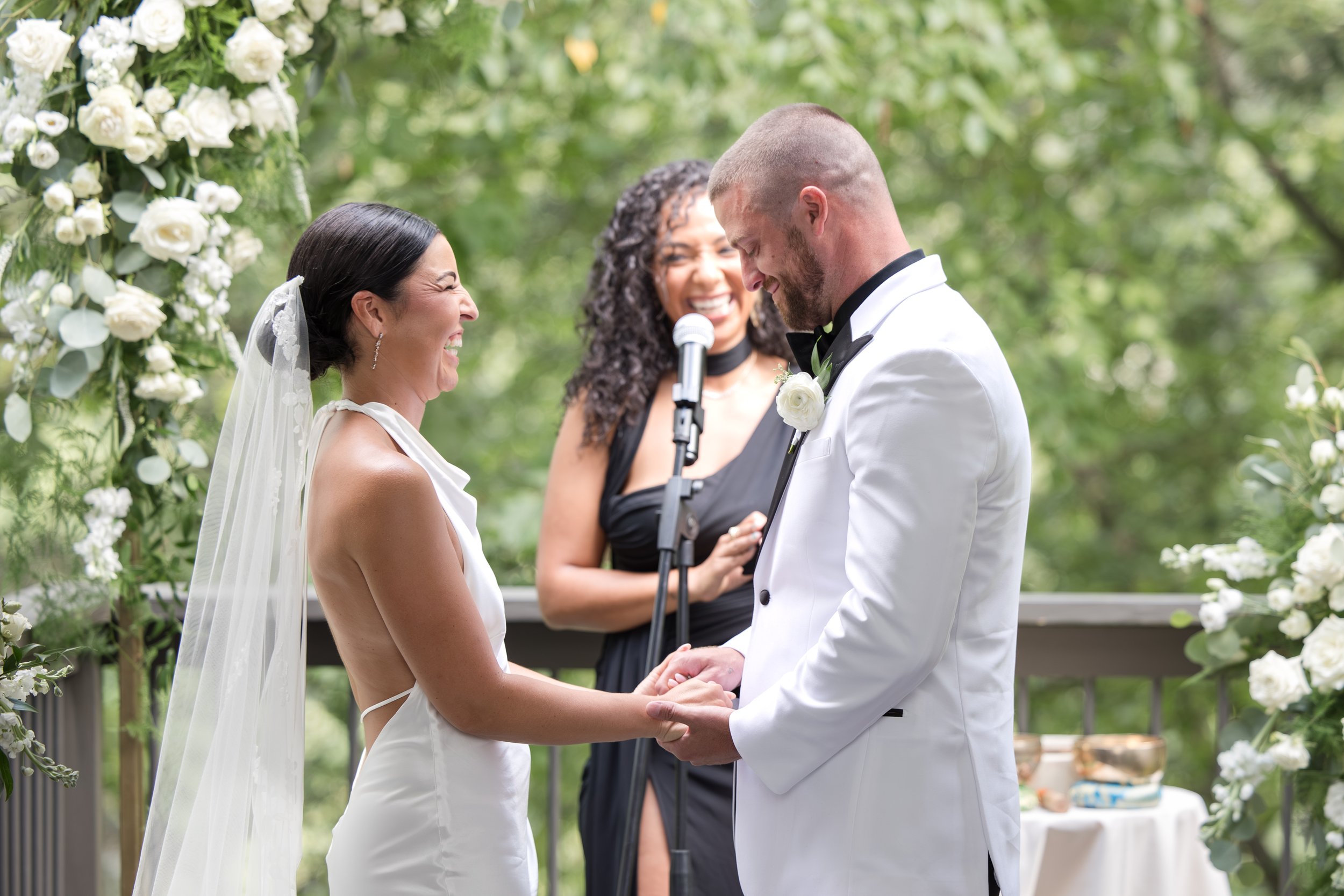 Bride and groom holding hands during their wedding ceremony outdoors, with officiant in the background, surrounded by greenery and white flowers.