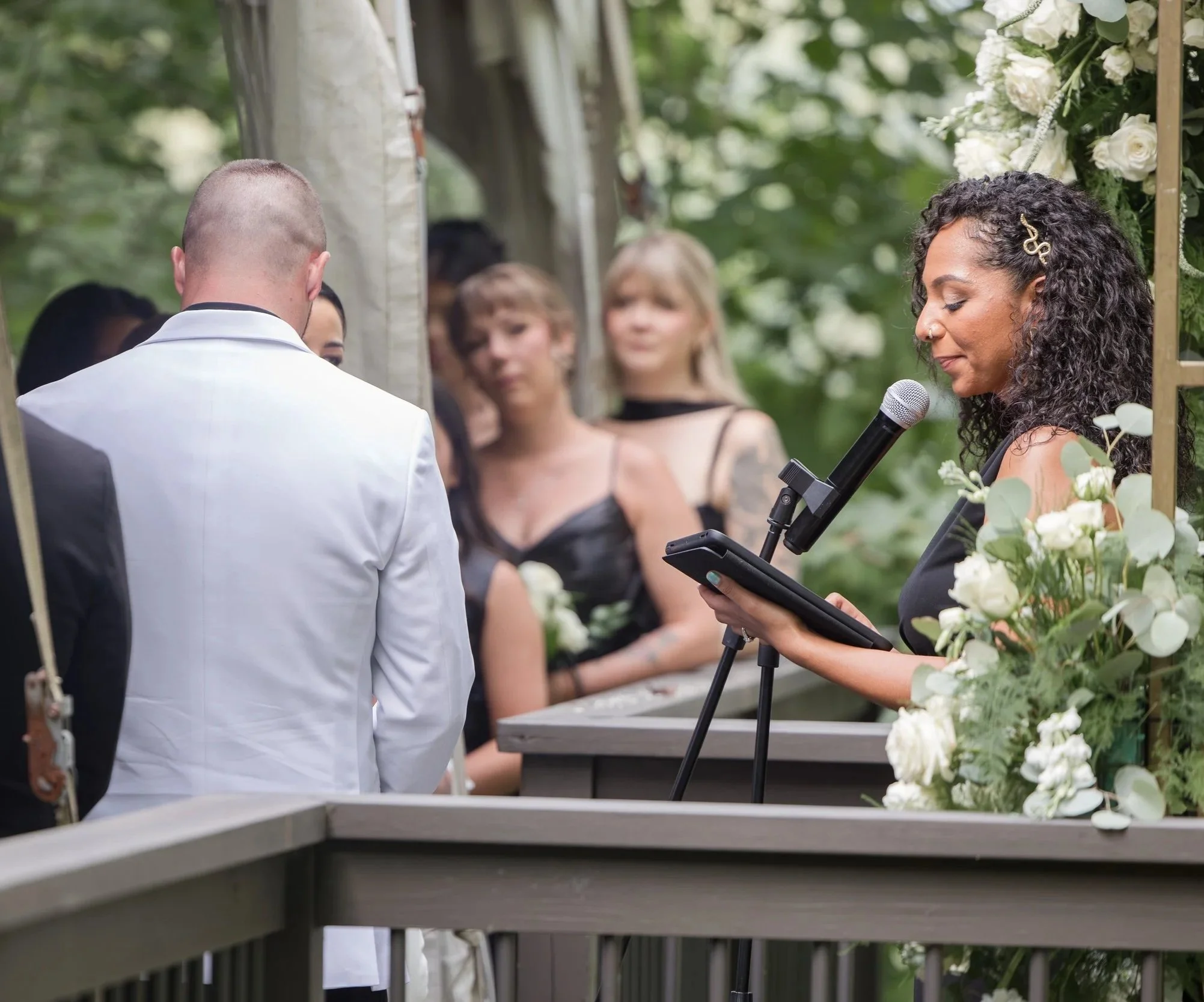 A woman officiant reading vows during an outdoor wedding ceremony with a bride and groom, and guests, in a natural setting.