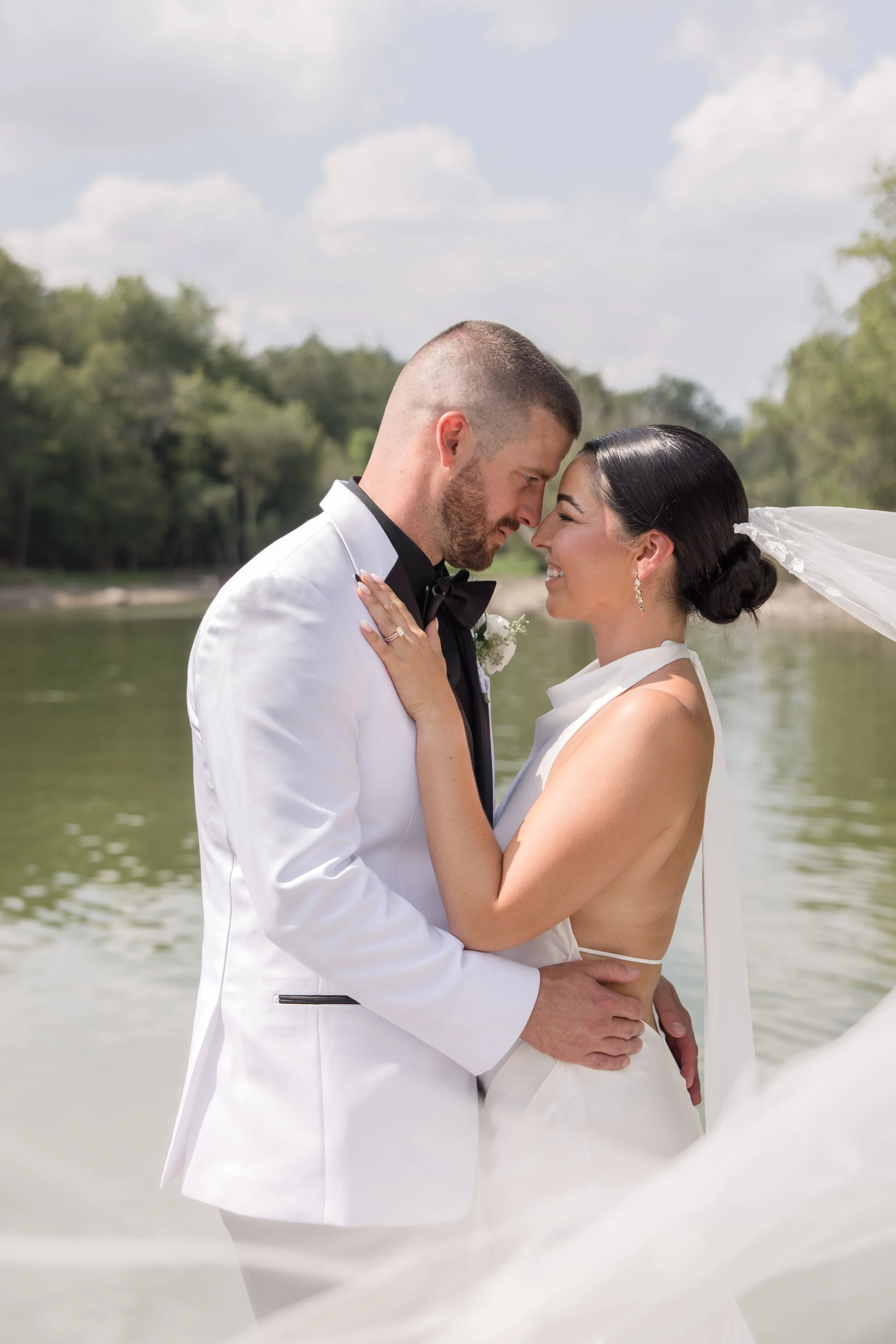 Couple in wedding attire sharing a close moment outdoors near a body of water, with trees and cloudy sky in the background.