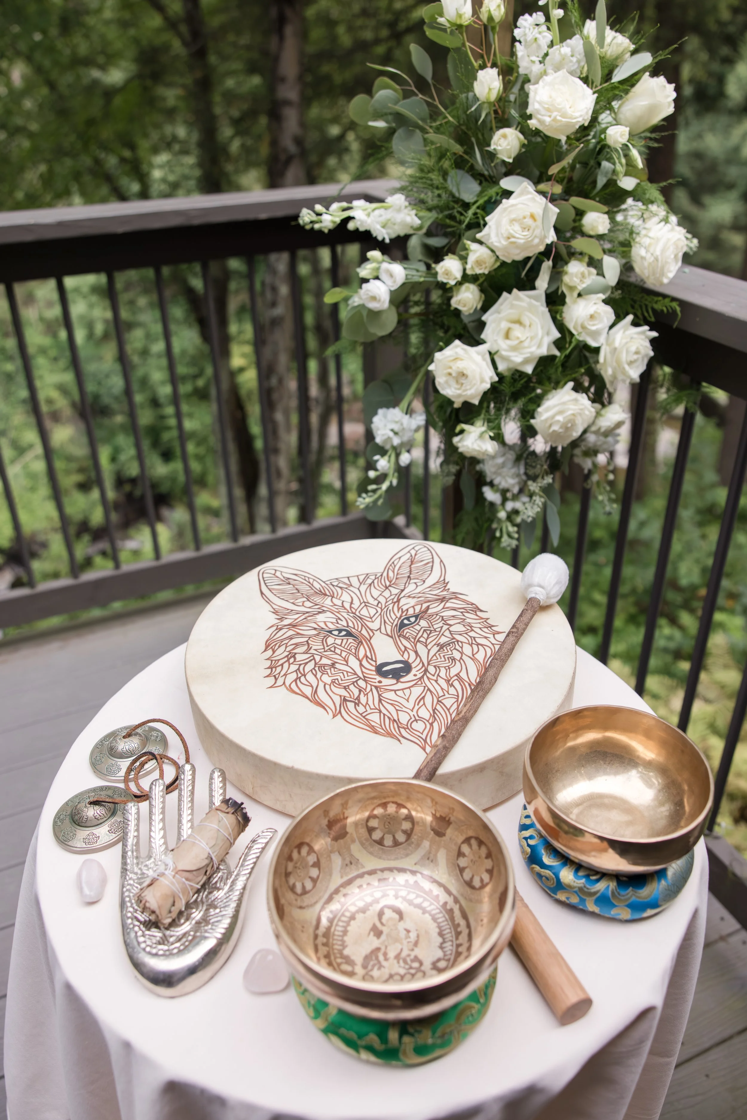 A table with a Christening set-up, including a round cake with a wolf illustration, singing bowls, a white cloth, white and copper hanging jewelry, a rolled scroll tied with string, and a white flower petal on a white tablecloth, outdoors on a wooden deck with green foliage and a floral arrangement in the background.