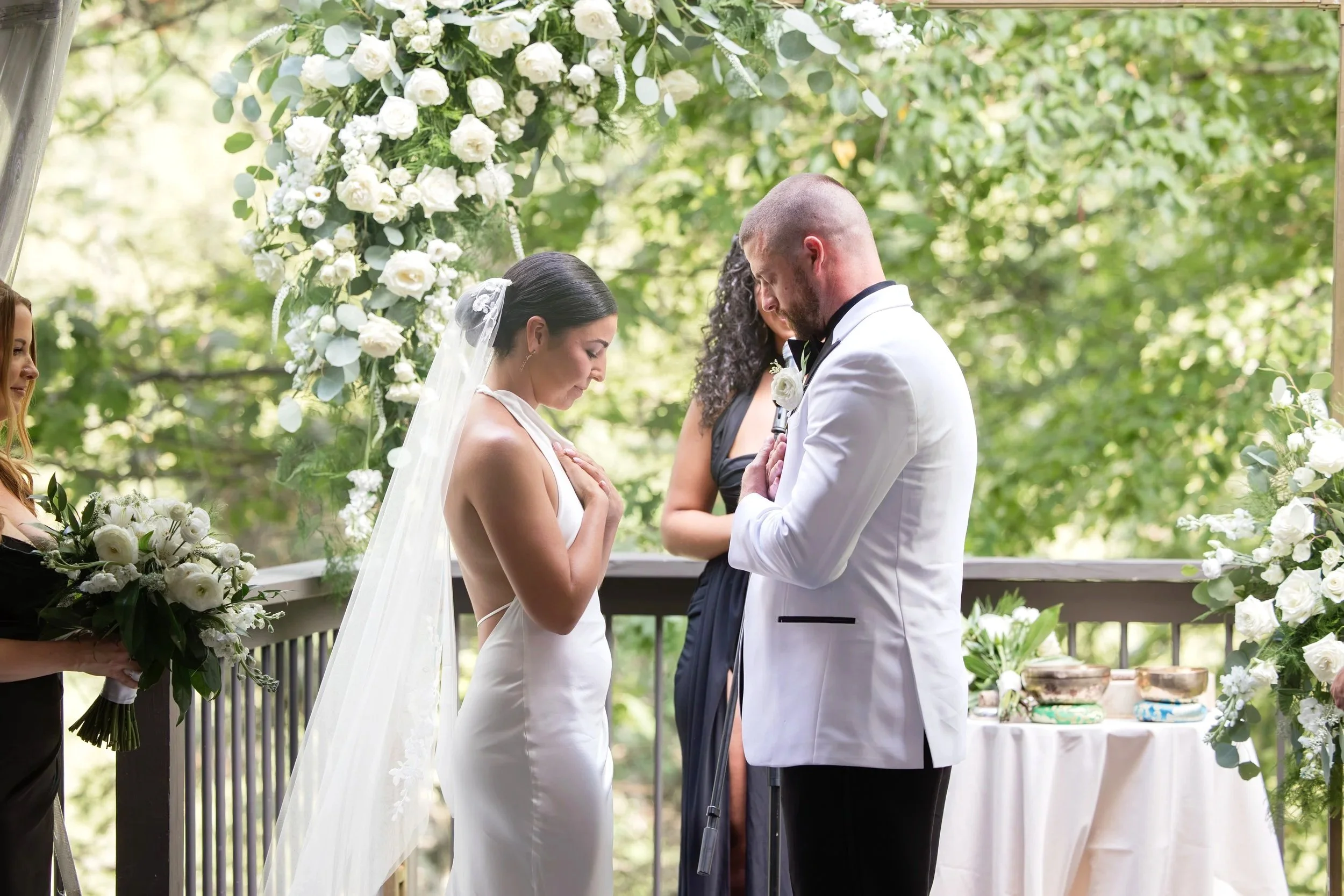 A wedding ceremony outdoors with a bride and groom praying, standing under a floral archway, with bridesmaids holding bouquets nearby.