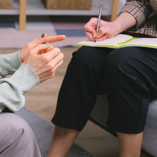 Two people having a conversation, one holding a pen and notebook, the other gesturing with hands.