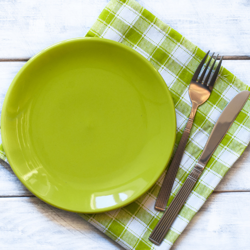 Empty green plate on a green and white checkered napkin with a fork and knife on a white wooden surface.