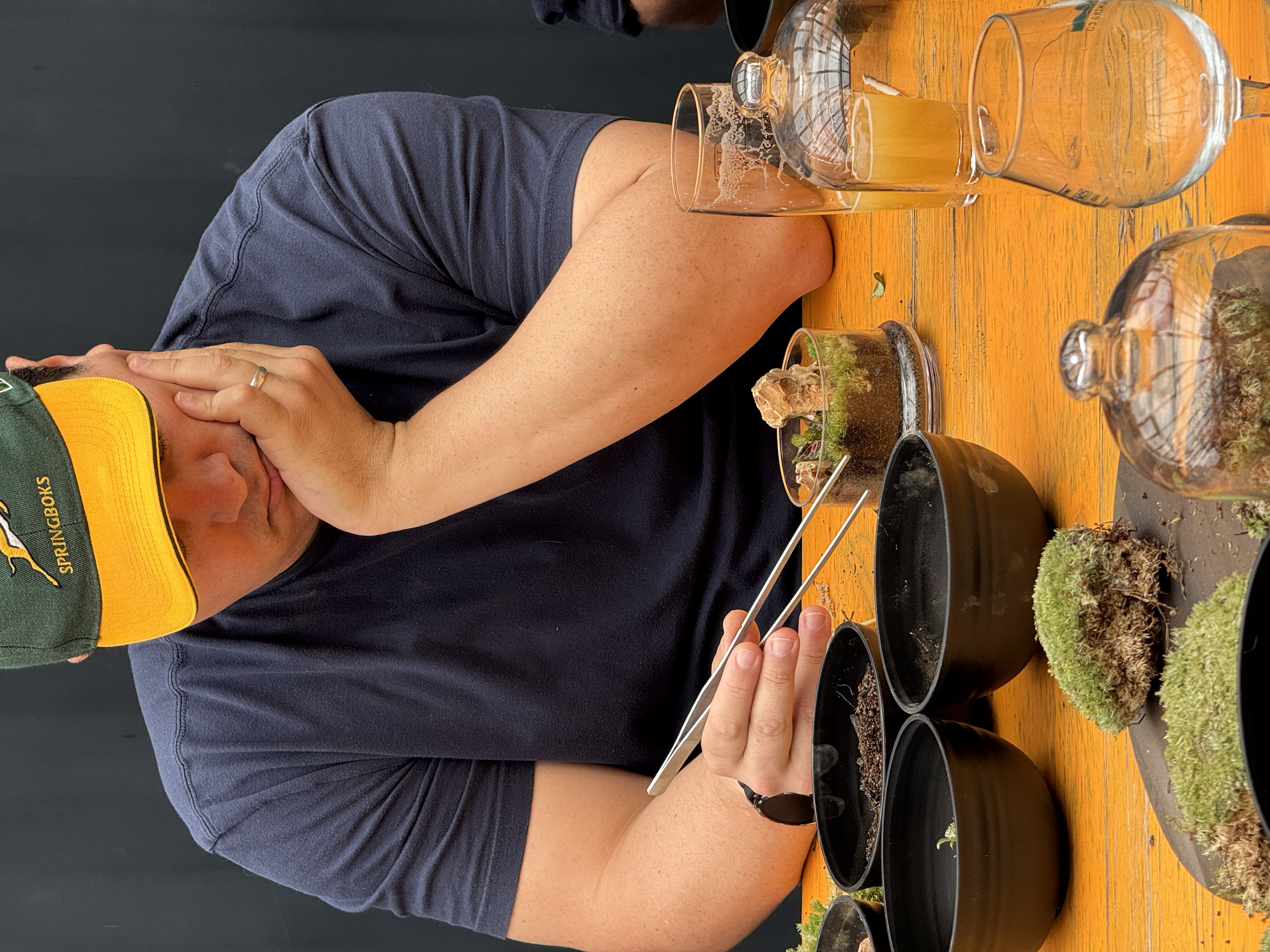 A man wearing a green and yellow baseball cap and a black t-shirt sitting at a wooden table filled with small glass terrariums and containers of moss and plants, with his face covered by his hand.
