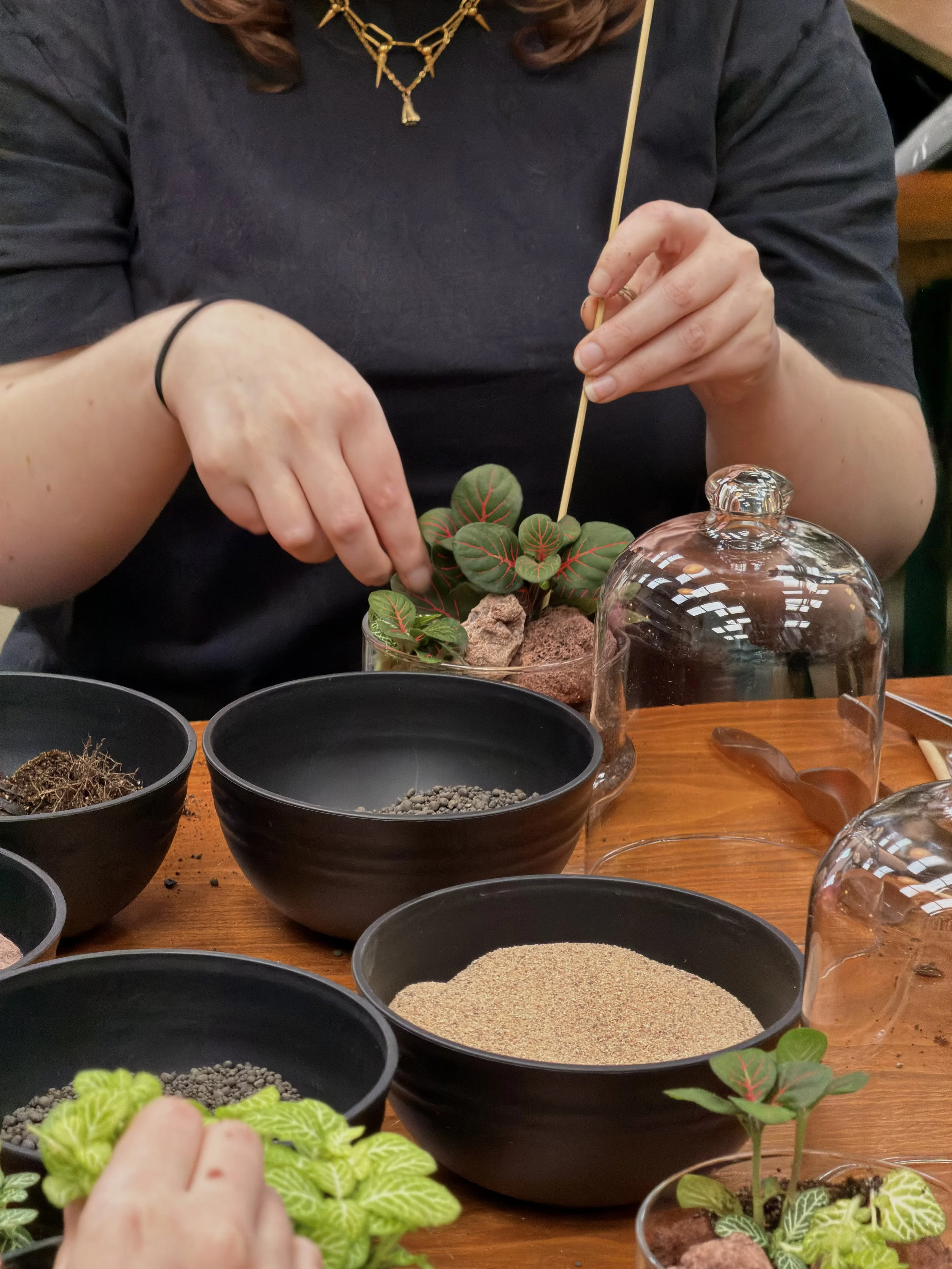 Person at a terrarium workshops arranging a potted plant with colourful leaves using a skewer, surrounded by bowls containing soil, small rocks, and sand, on a wooden table.