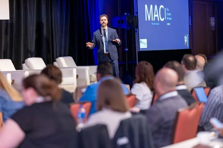 Speaker on stage at a conference, addressing seated audience, with "MAC The Creator Mindset" displayed on screen.