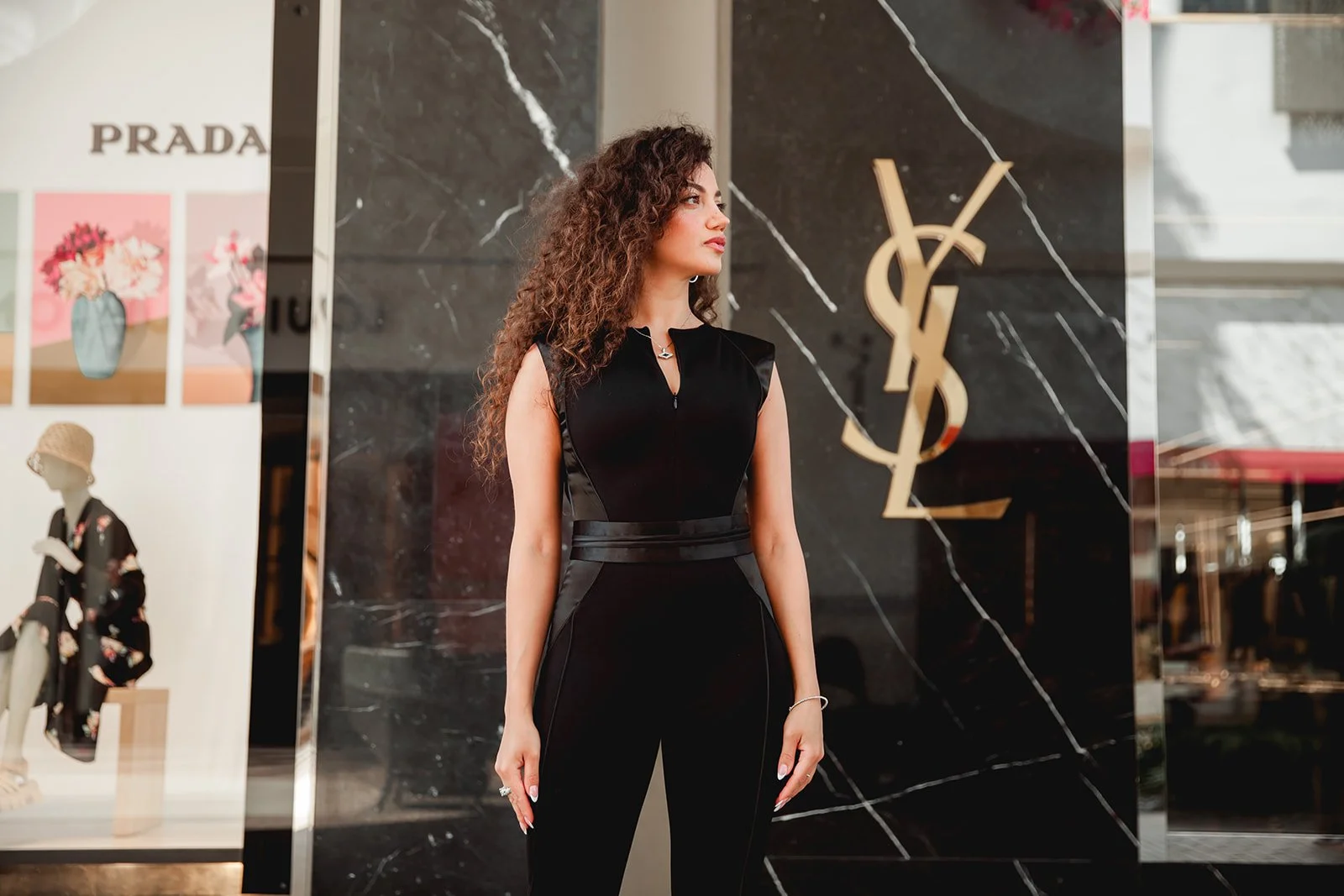 A woman stands in front of a store display, dressed in a black outfit. The backdrop has a Prada sign and YSL logo on a marble wall. A mannequin wearing floral attire and a hat is visible in the window display.