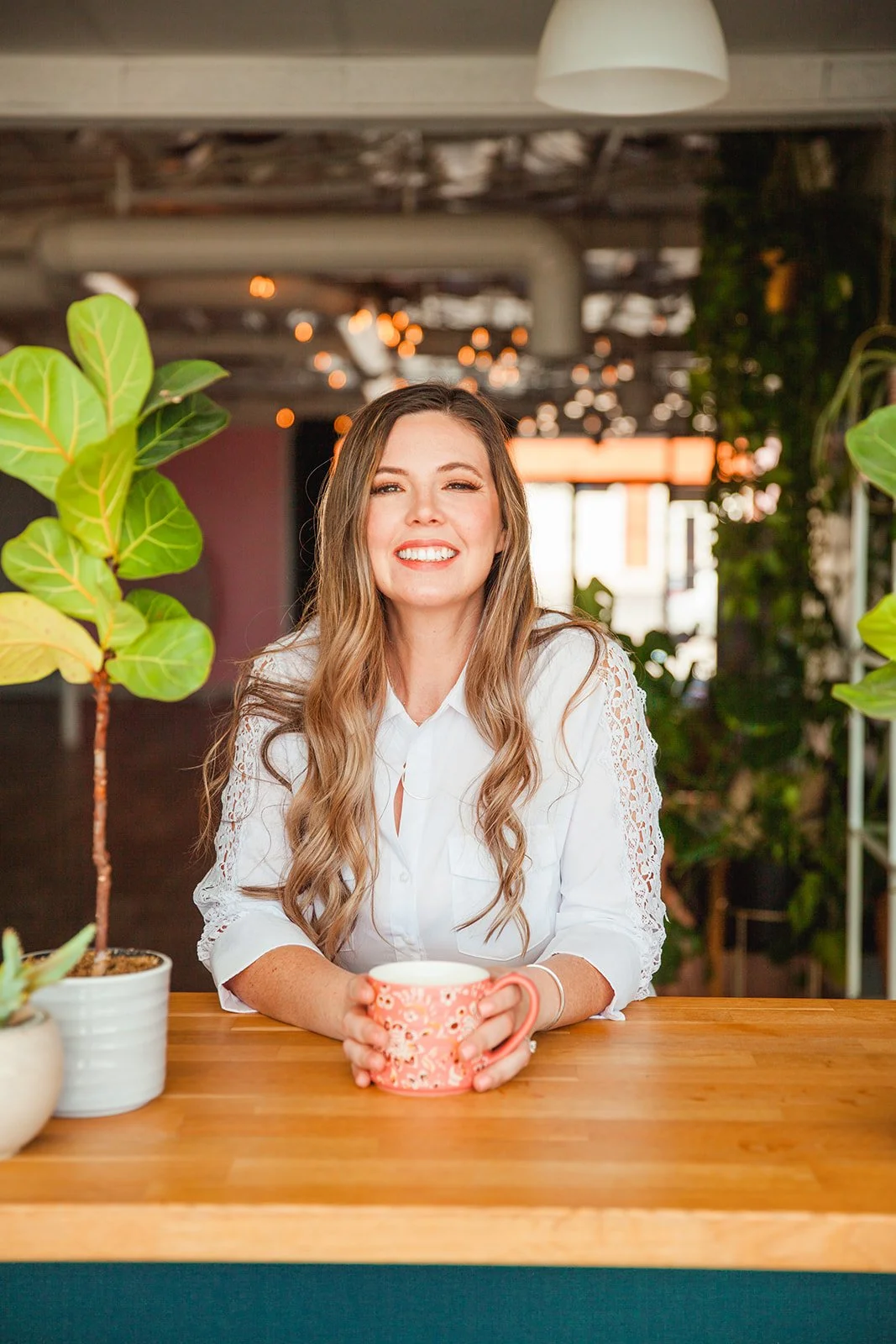 Smiling woman with long hair holding a mug, sitting at a wooden table with potted plants, in a modern indoor space.