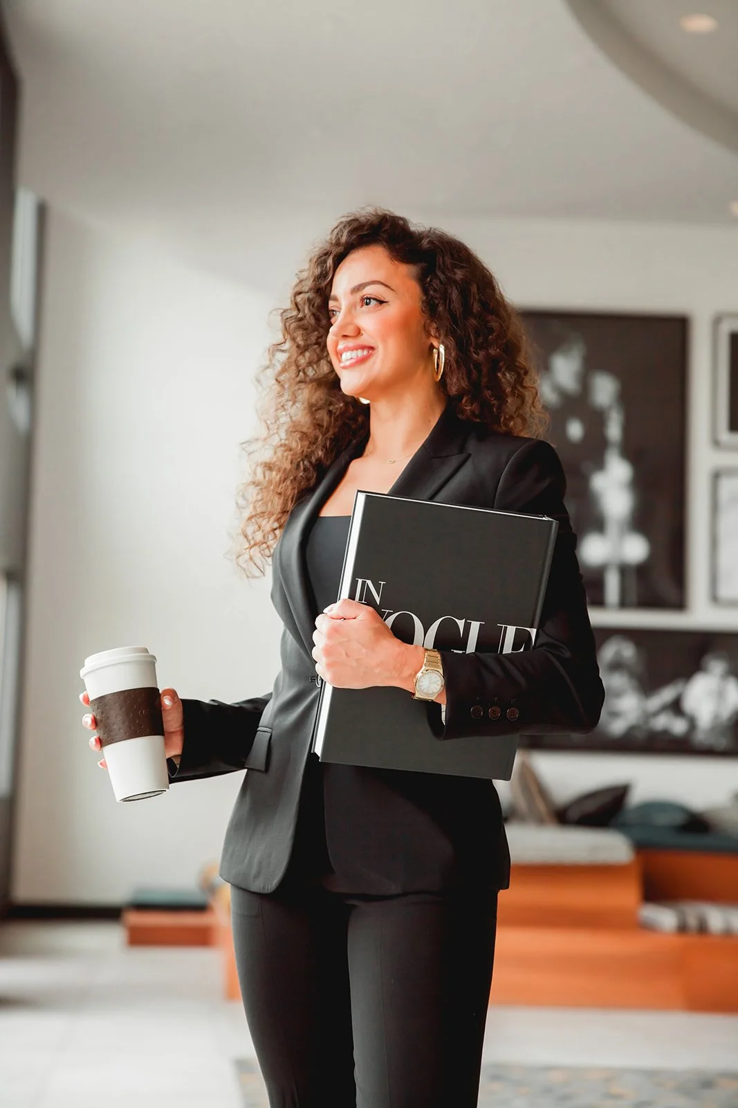 Smiling woman in business attire holding a coffee cup and a magazine in a modern office setting.