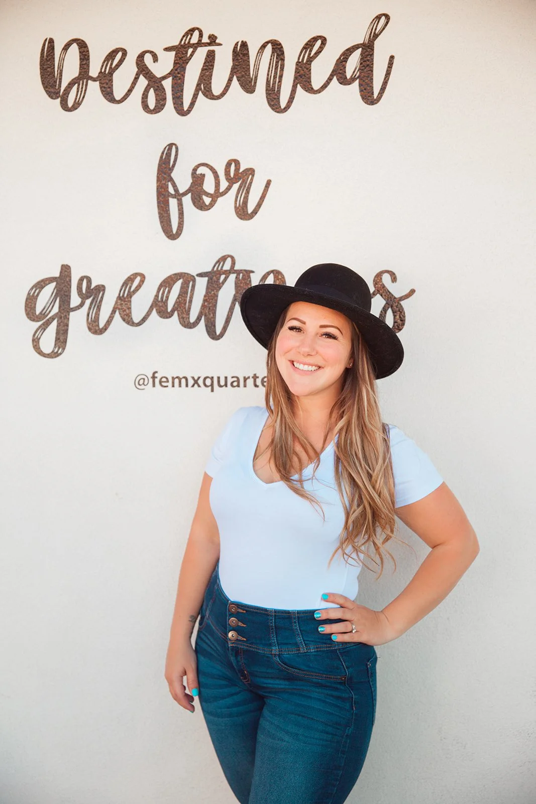 Woman wearing black hat and jeans standing in front of sign reading 'Destined for greatness' with Instagram handle @femxquarter.