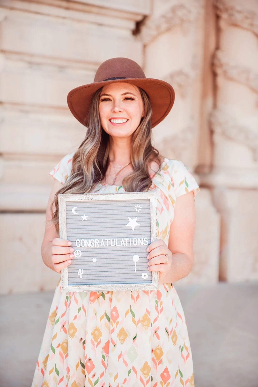 Woman smiling, wearing a brown hat and patterned dress, holding a sign with the word "Congratulations" and decorative icons.